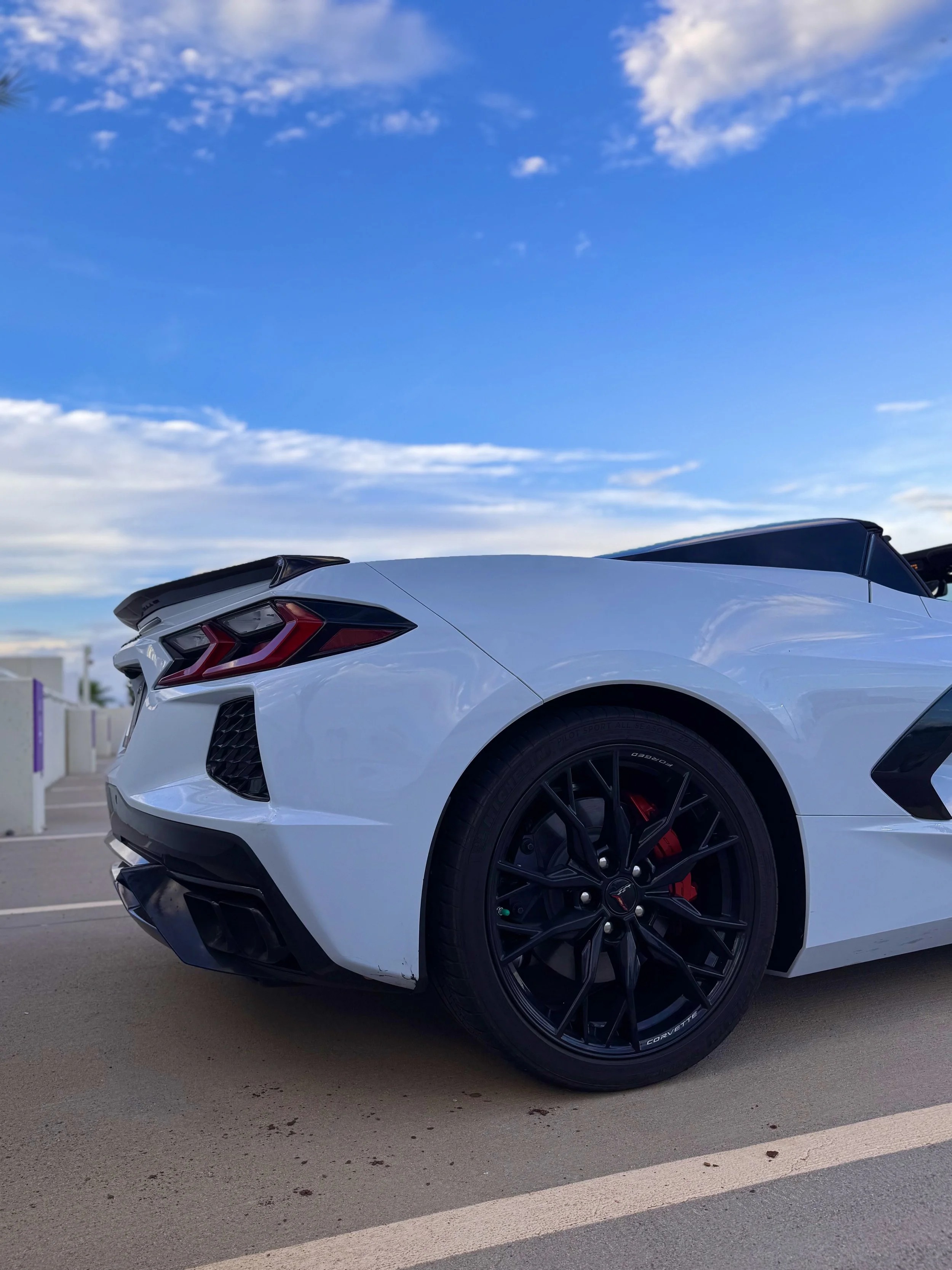 A White Chevrolet Corvette C8 convertible sports car parked on a rooftop parking lot under a blue sky with clouds, featuring black wheels and red brake calipers.