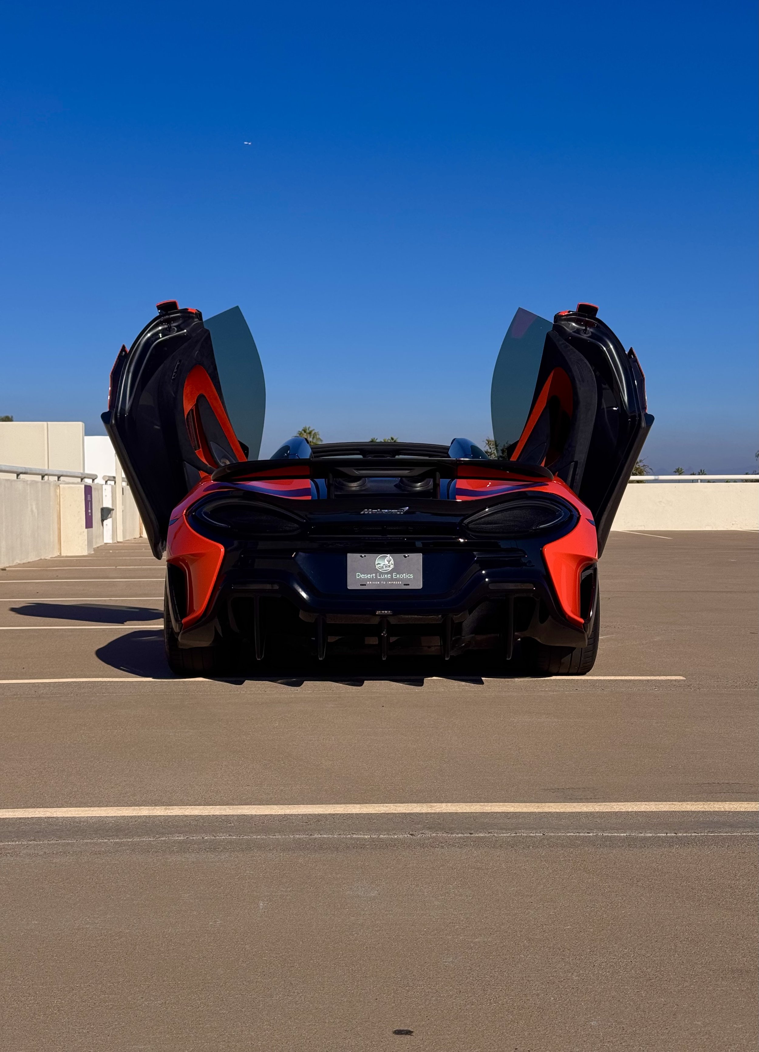 A tuned and straight-piped Helios orange McLaren 600LT spider exotic sports car with upward opening butterfly doors parked in an empty parking lot, blue sky in the background.