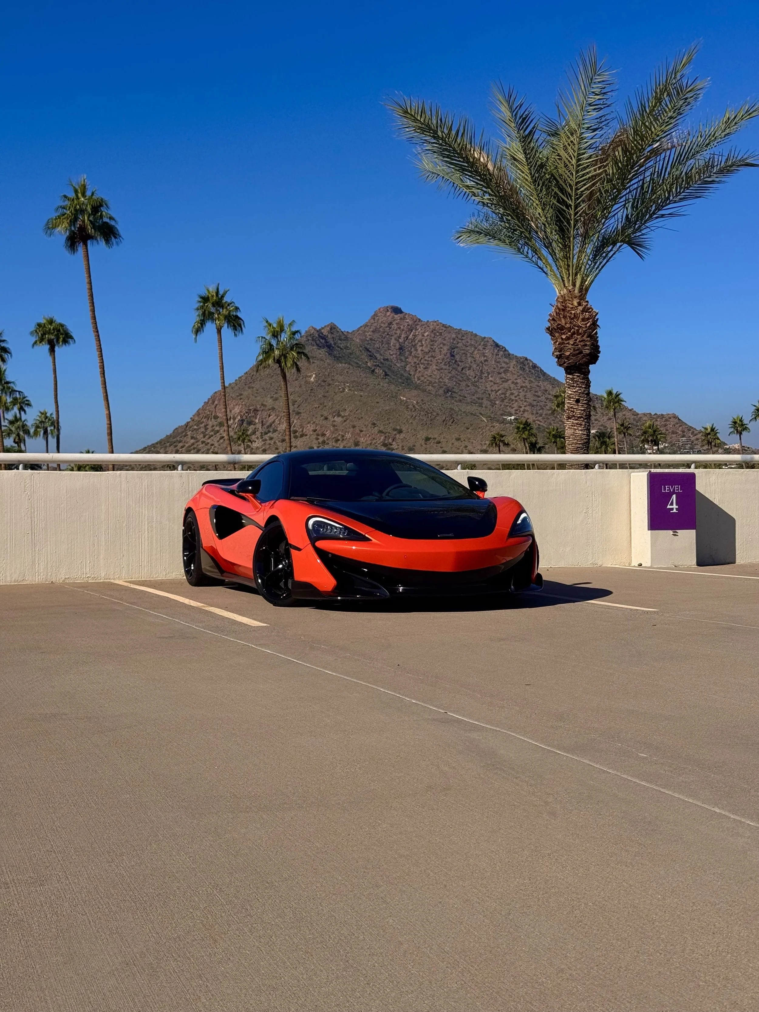 A tuned and straight-piped Helios orange McLaren 600LT spider exotic sports car parked in an outdoor lot with palm trees, a mountain, and a clear blue sky in the background.