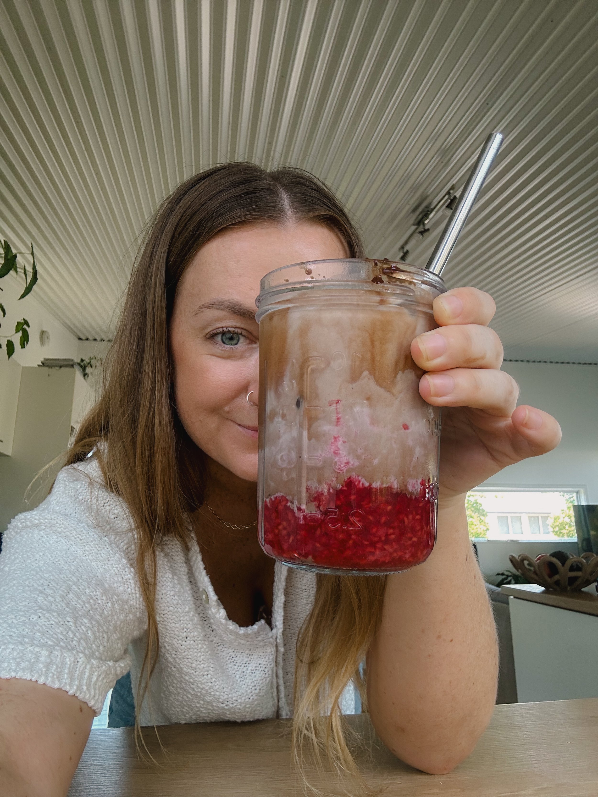 A woman with long brown hair, wearing a white textured top, holding a large glass jar with a metal straw, filled with a drink that has red berries at the bottom and a creamy, frothy top, sitting in a kitchen with a wooden table.