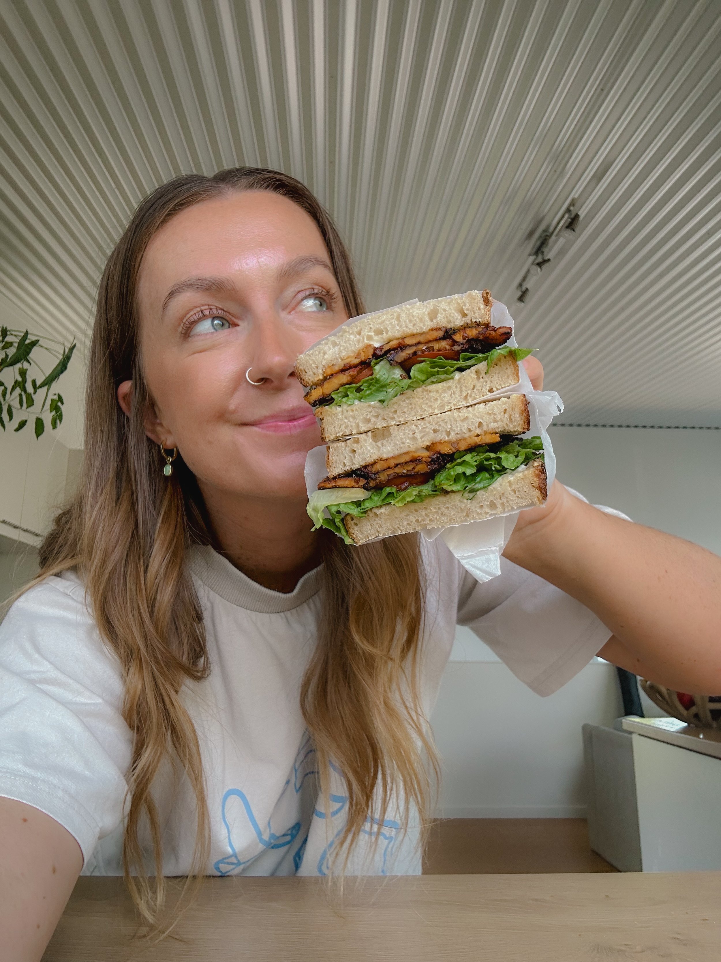 Woman with long wavy hair, light skin, and a nose ring holding a large triple-decker sandwich near her face, smiling and looking away. The sandwich contains lettuce, tomato, and meat between slices of white bread.