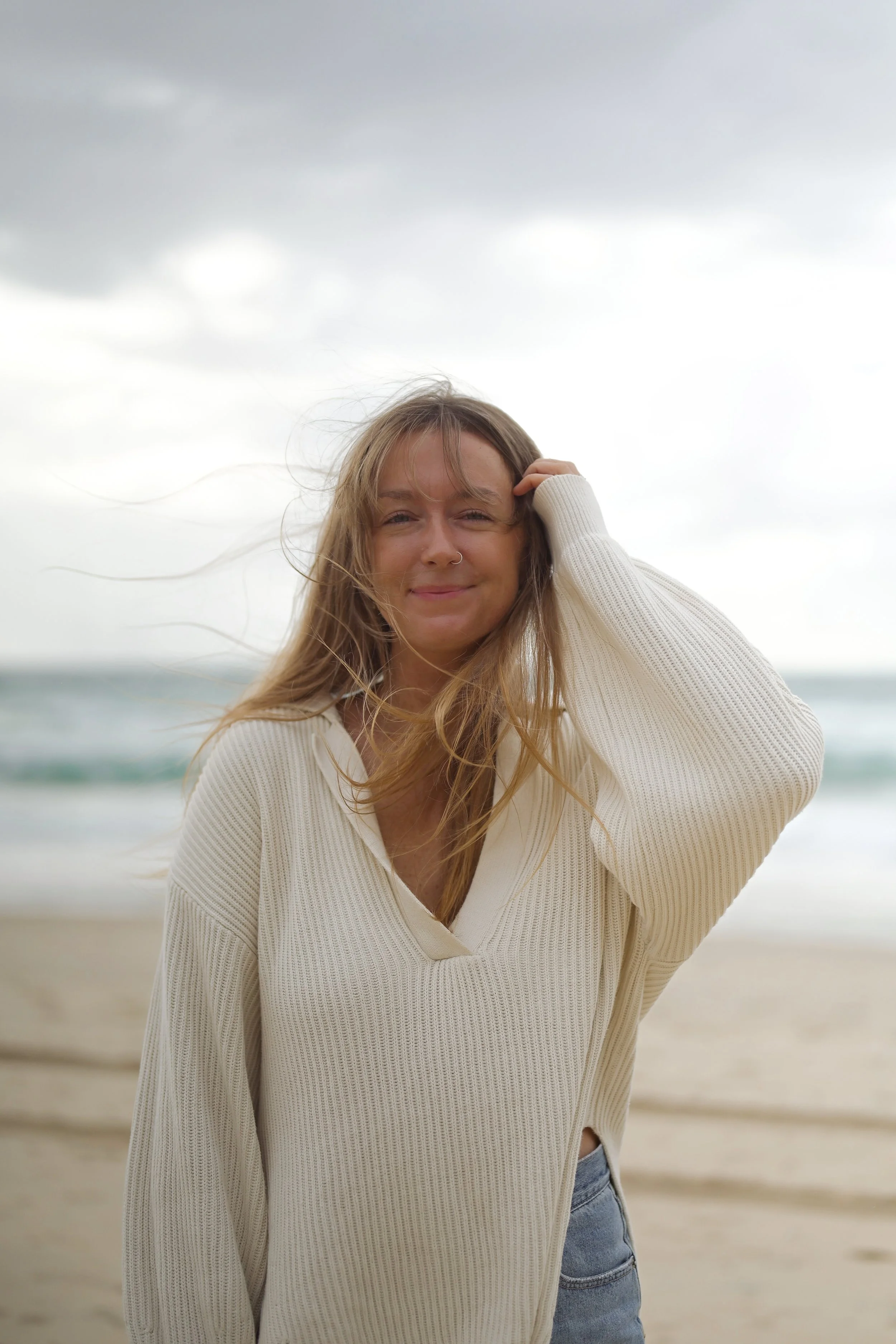 Woman with long hair smiling on a beach with cloudy sky, wearing a cream sweater and jeans.