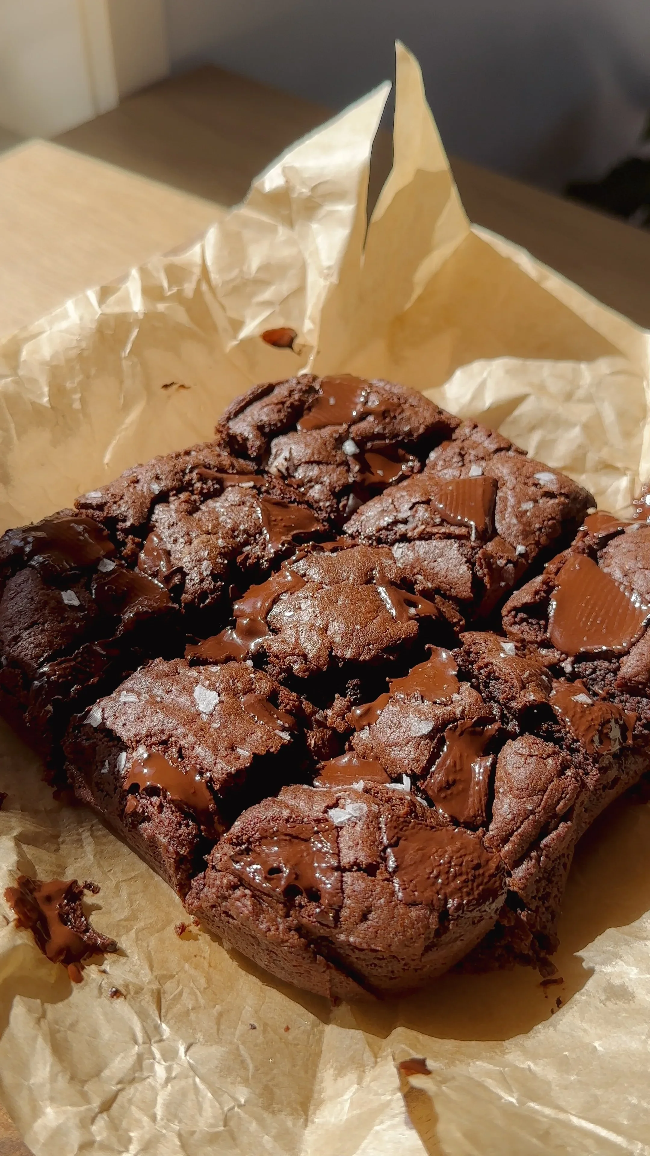 A close-up of a chocolate brownies with chocolate pieces on top, placed on crumpled parchment paper.