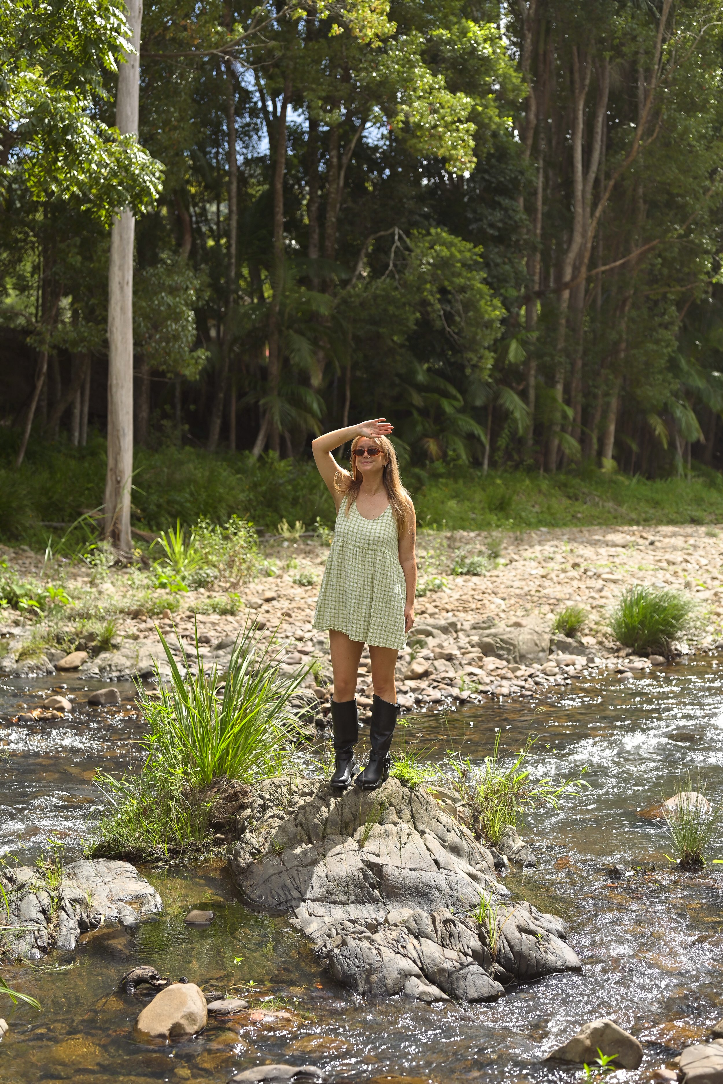 A woman standing on a large rock in a shallow creek, shielding her eyes from the sun with her hand, in a wooded outdoor setting.