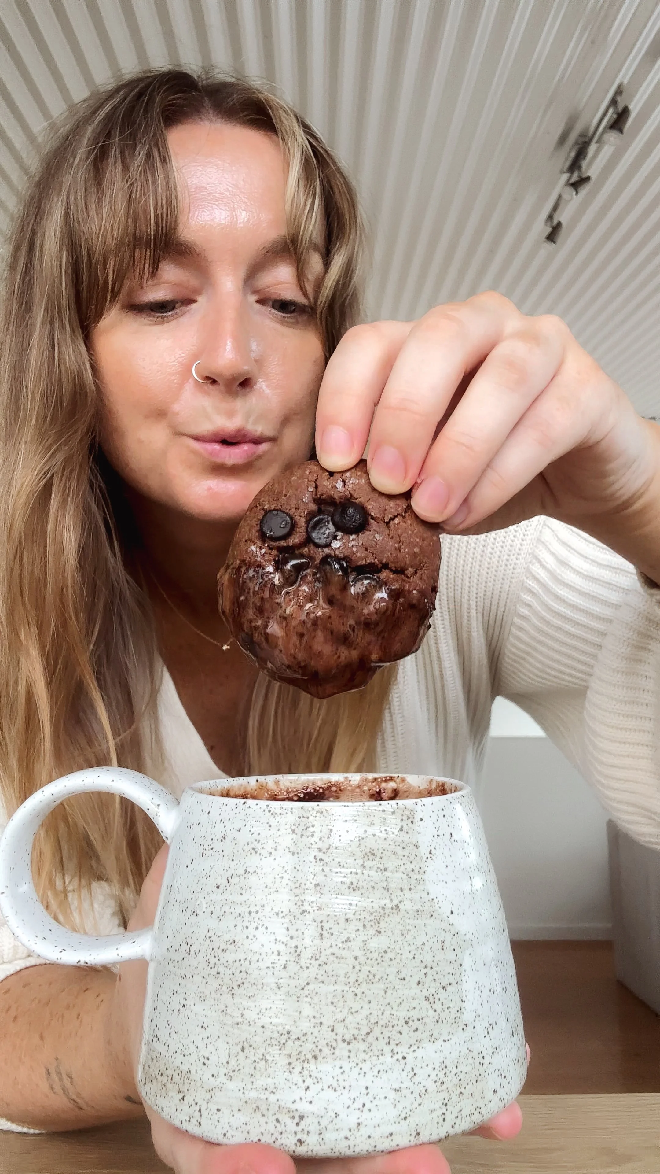 A woman with long blonde hair and a nose ring holds a large chocolate cookie above a white speckled mug, appearing to dunk or dip it into a hot chocolate or similar beverage.
