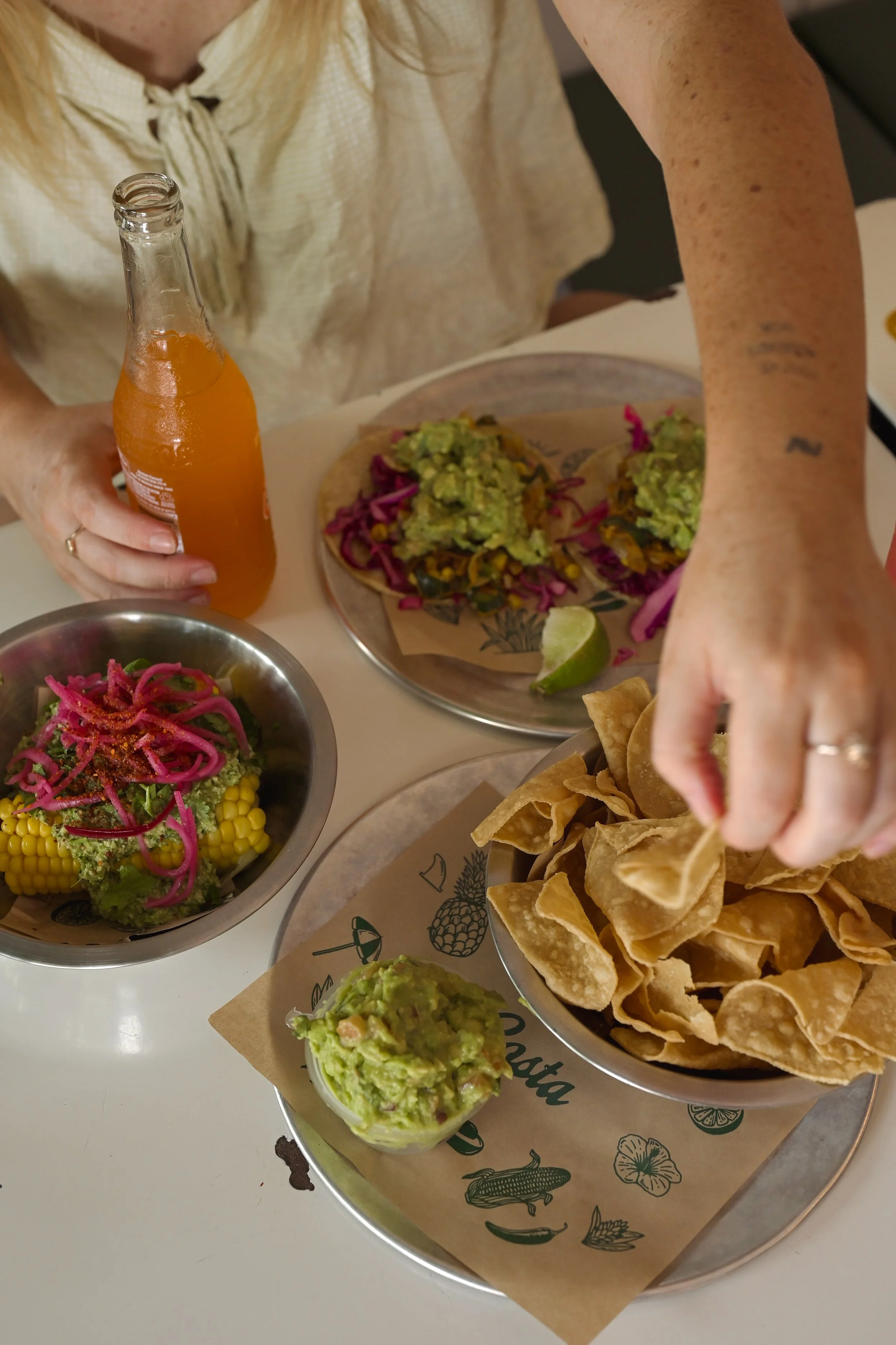 A person reaching for tortilla chips with guacamole on a tray, with bowls of salad, tacos, and a bottle of orange soda on the table.