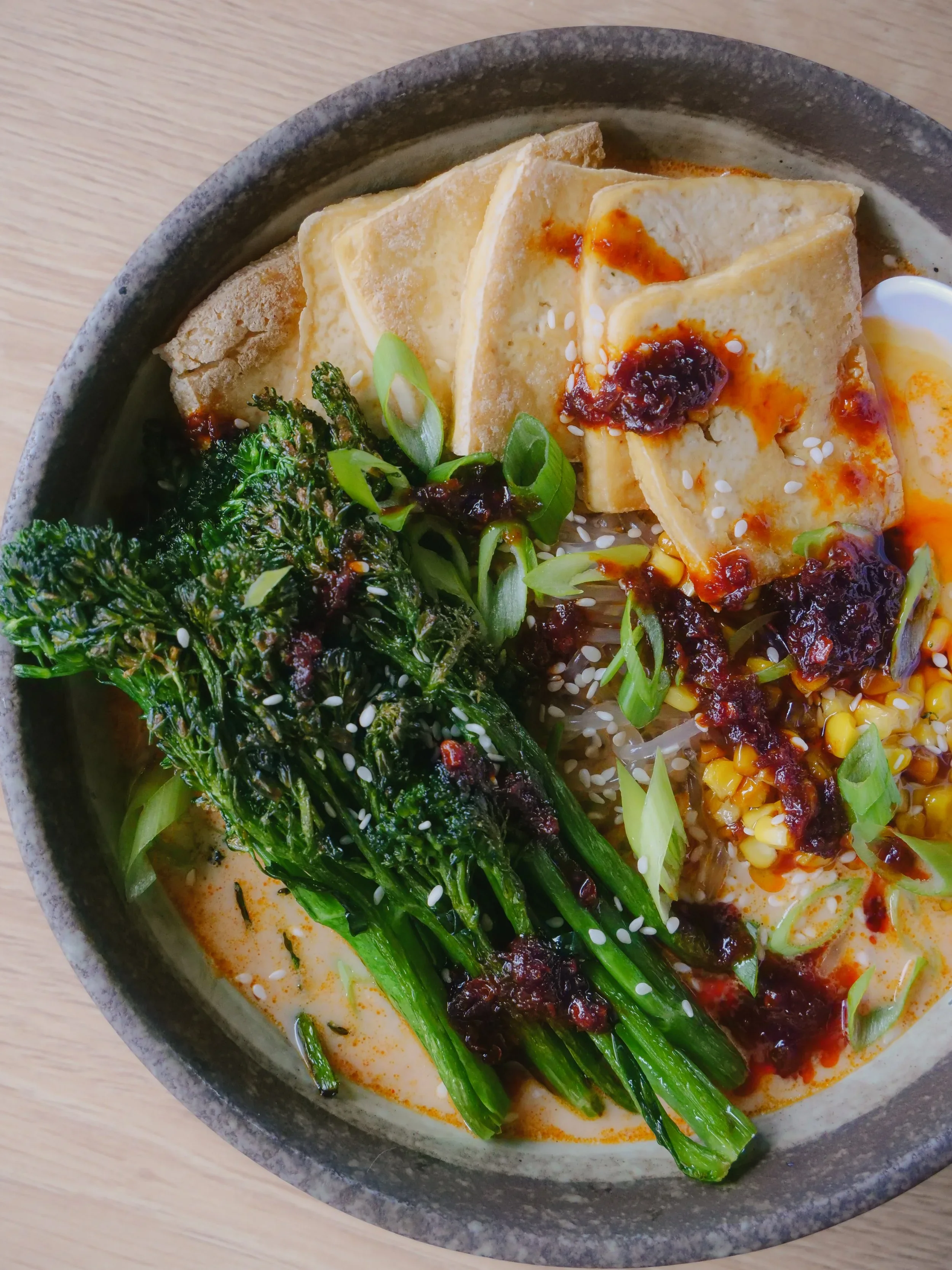 A bowl of ramen with green leafy vegetables, tofu topped with sauce, chopped green onions, sesame seeds, and a soft boiled egg in a flavorful broth.