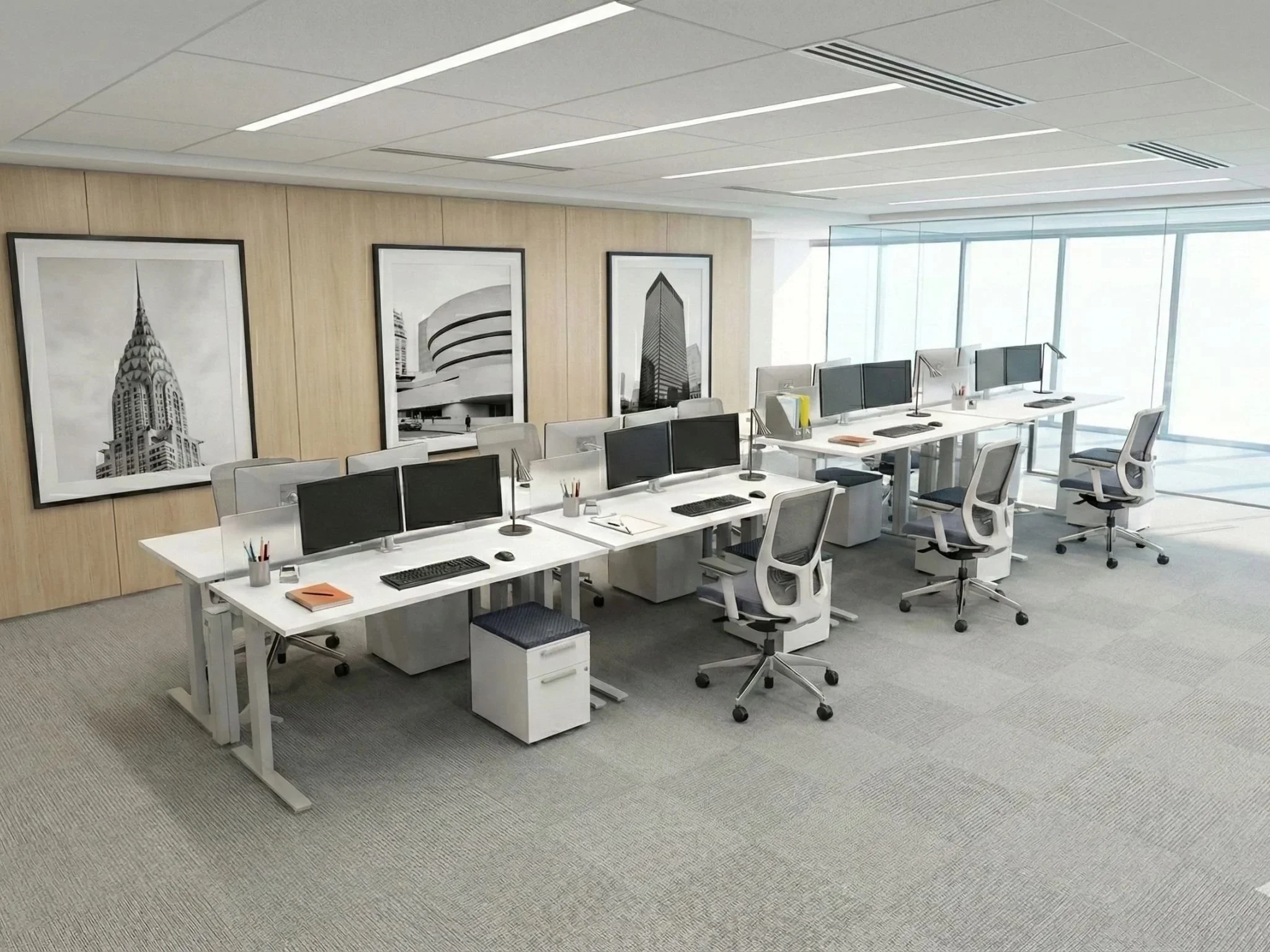 An office with white desks and chairs arranged in a row, each desk equipped with computer monitors, keyboards, mice, and office supplies. Three framed black-and-white architectural photographs hang on a beige wooden wall. Large windows on the right side allow natural light into the room.