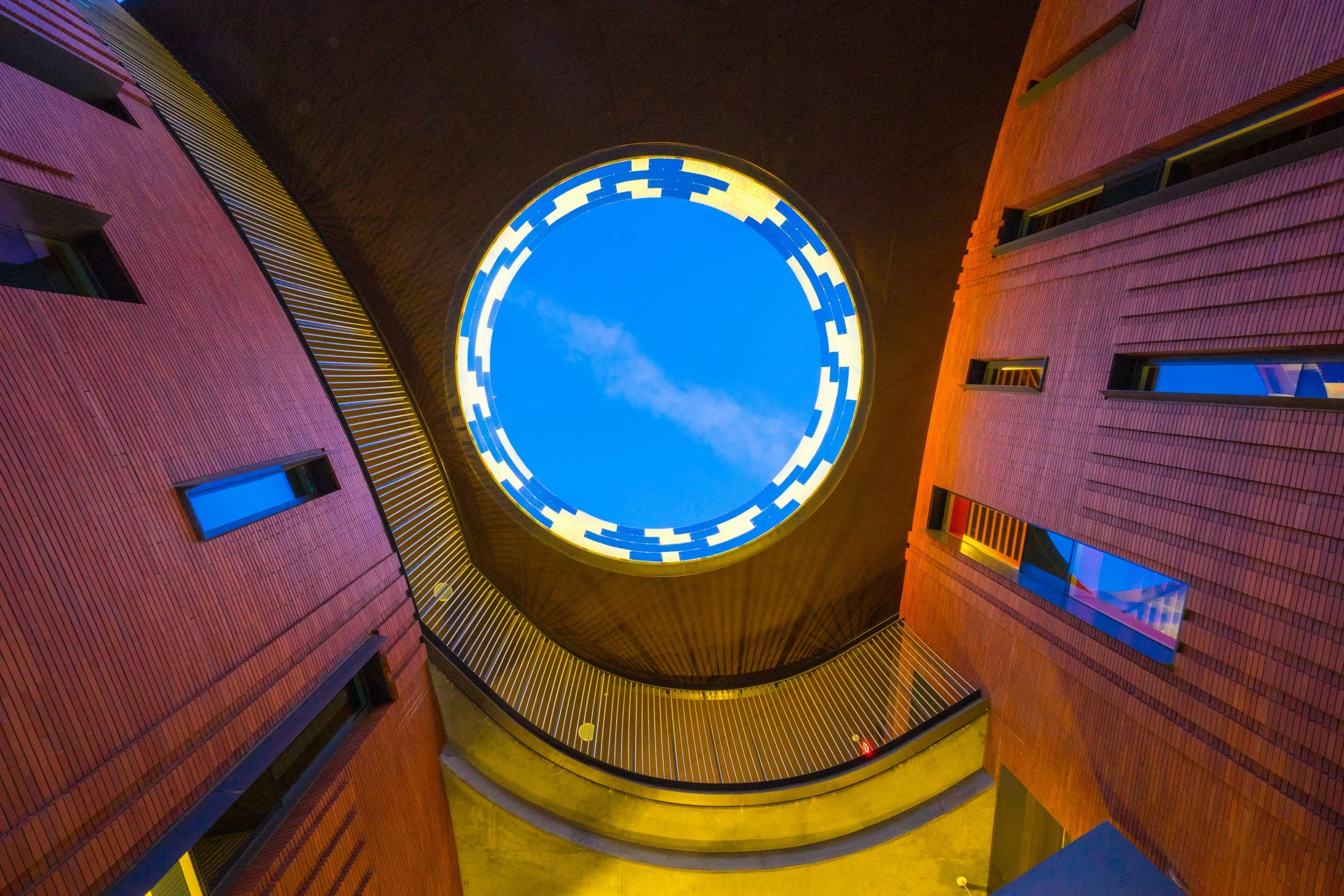 Interior view looking up at a circular skylight in a building with orange brick walls and large windows.