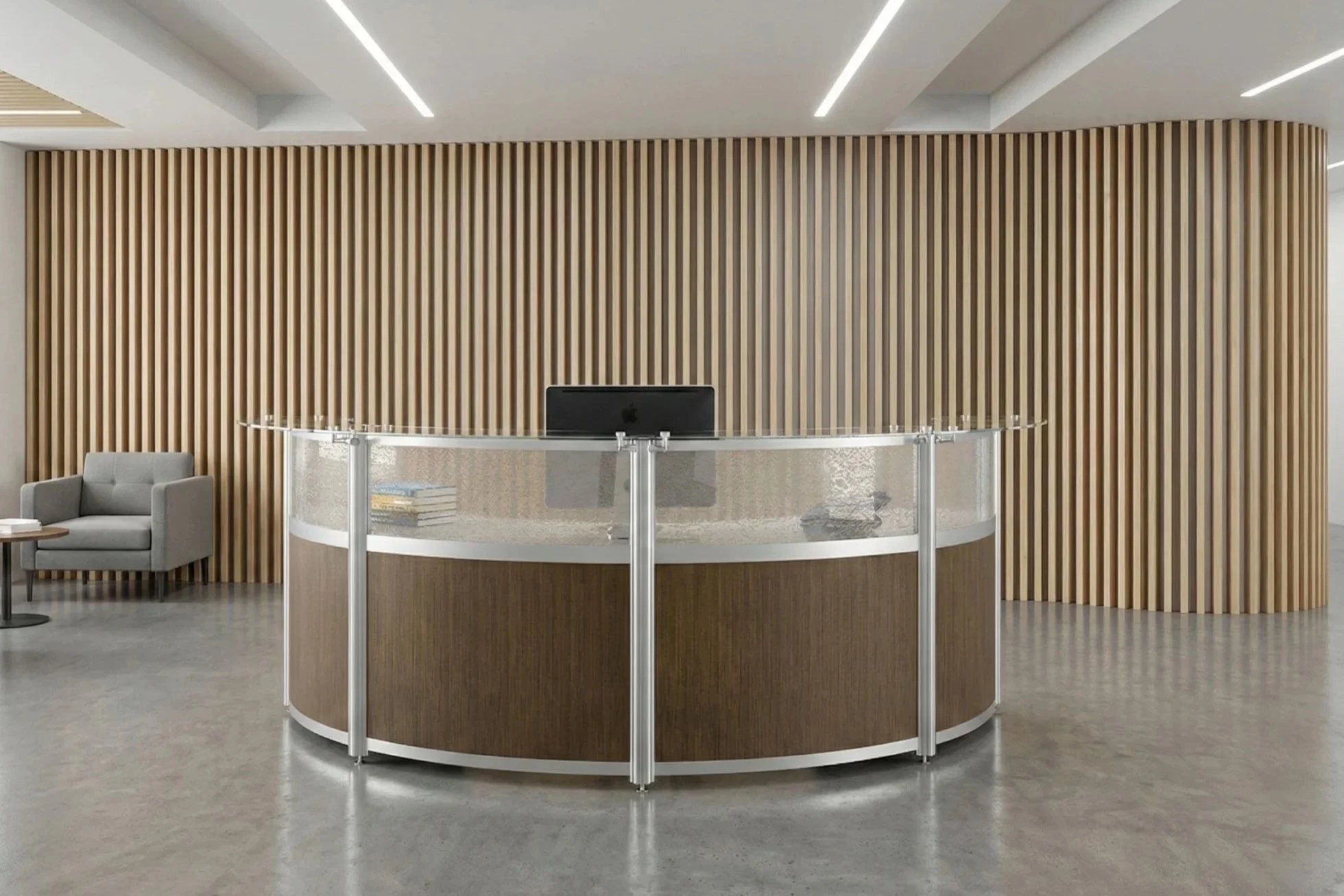 Modern reception area with a semi-circular wooden front desk, a black computer monitor, gray armchair, and a small side table with books. Behind the desk is a curved wooden slat wall, with a drop ceiling featuring linear recessed lighting.