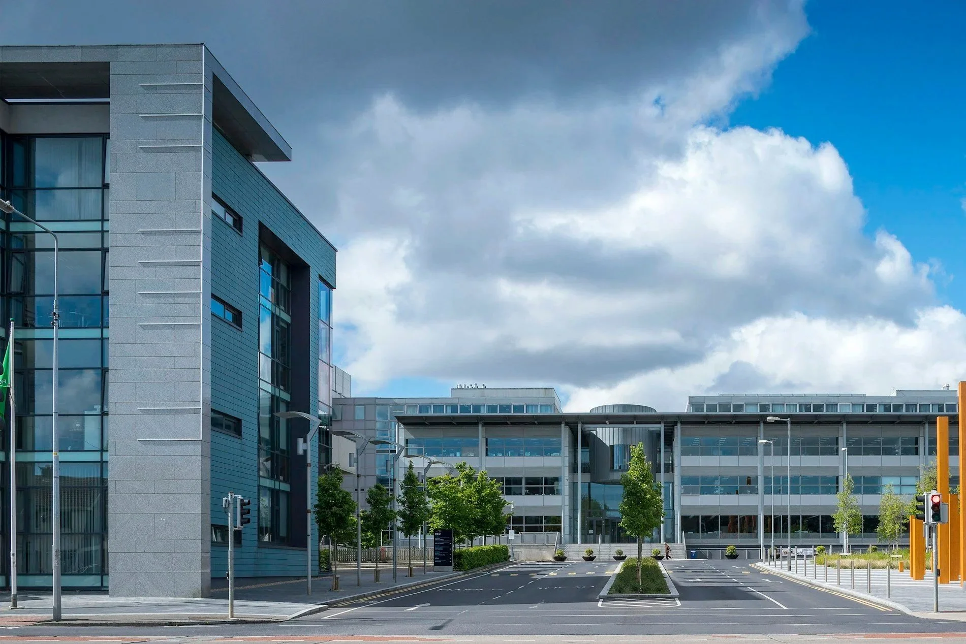 Modern office buildings with glass and metal facades in an urban setting, cloudy sky overhead, and a few green trees and street signs in the foreground.