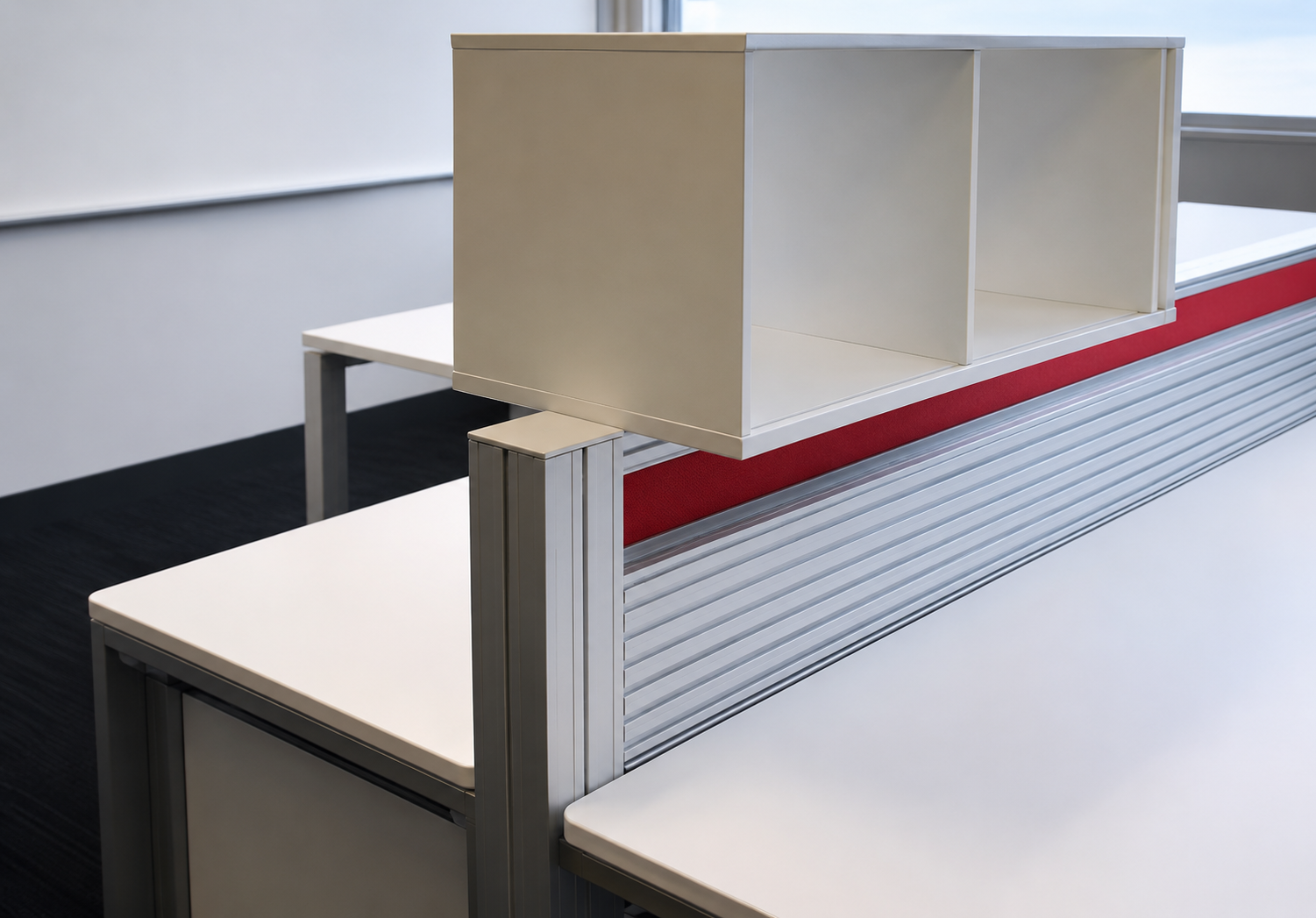 Empty office cubicle with white desk, beige partition, and red fabric separator, located near a window.
