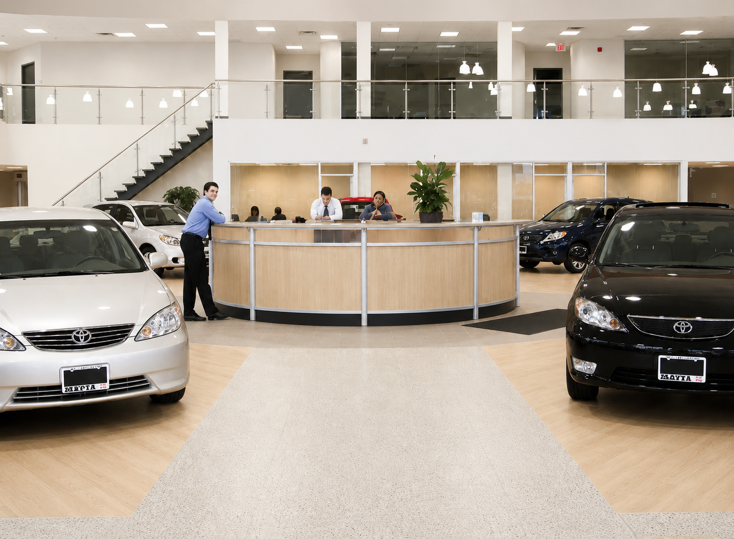 Car dealership showroom with a white and a black Toyota parked in front of a reception desk. Two employees behind the desk are working, with one person leaning on the desk and another talking on the phone. There are additional cars displayed behind the desk, and a staircase leading to the upper level.