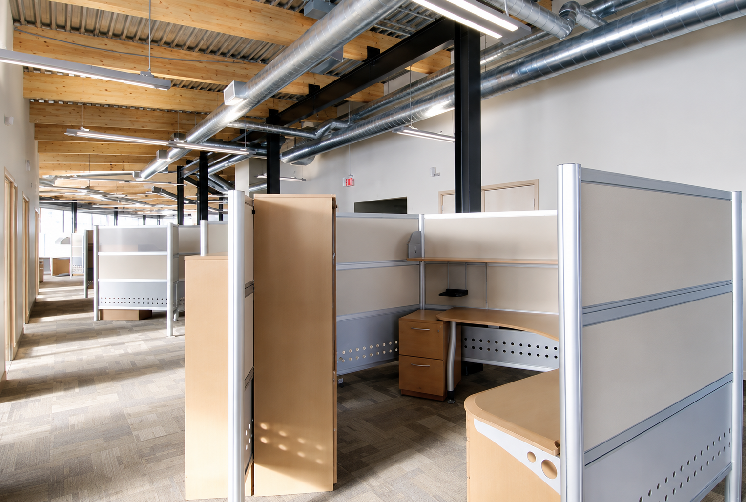 Empty office cubicles with wooden desks and beige dividers in a modern workspace with exposed ductwork and wooden ceiling beams.