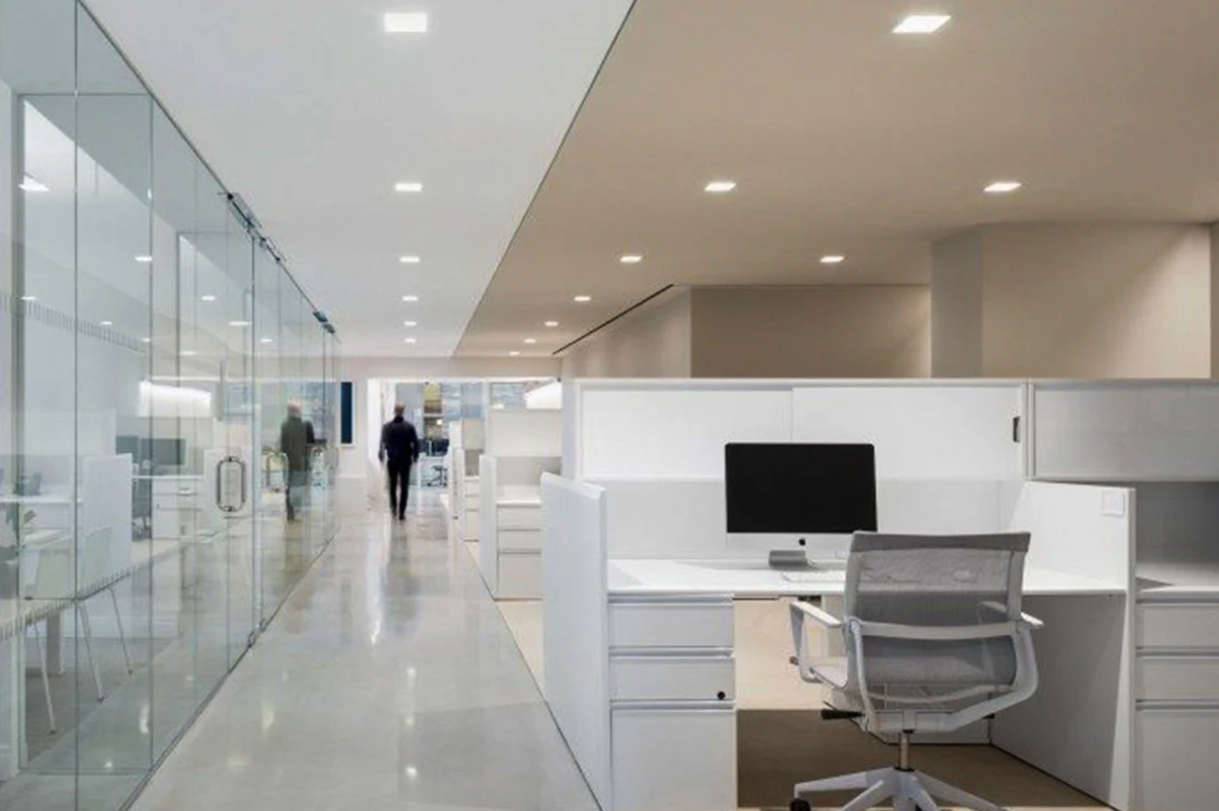 Modern office with arranged cubicles, a computer on the desk, and two people walking in the hallway.