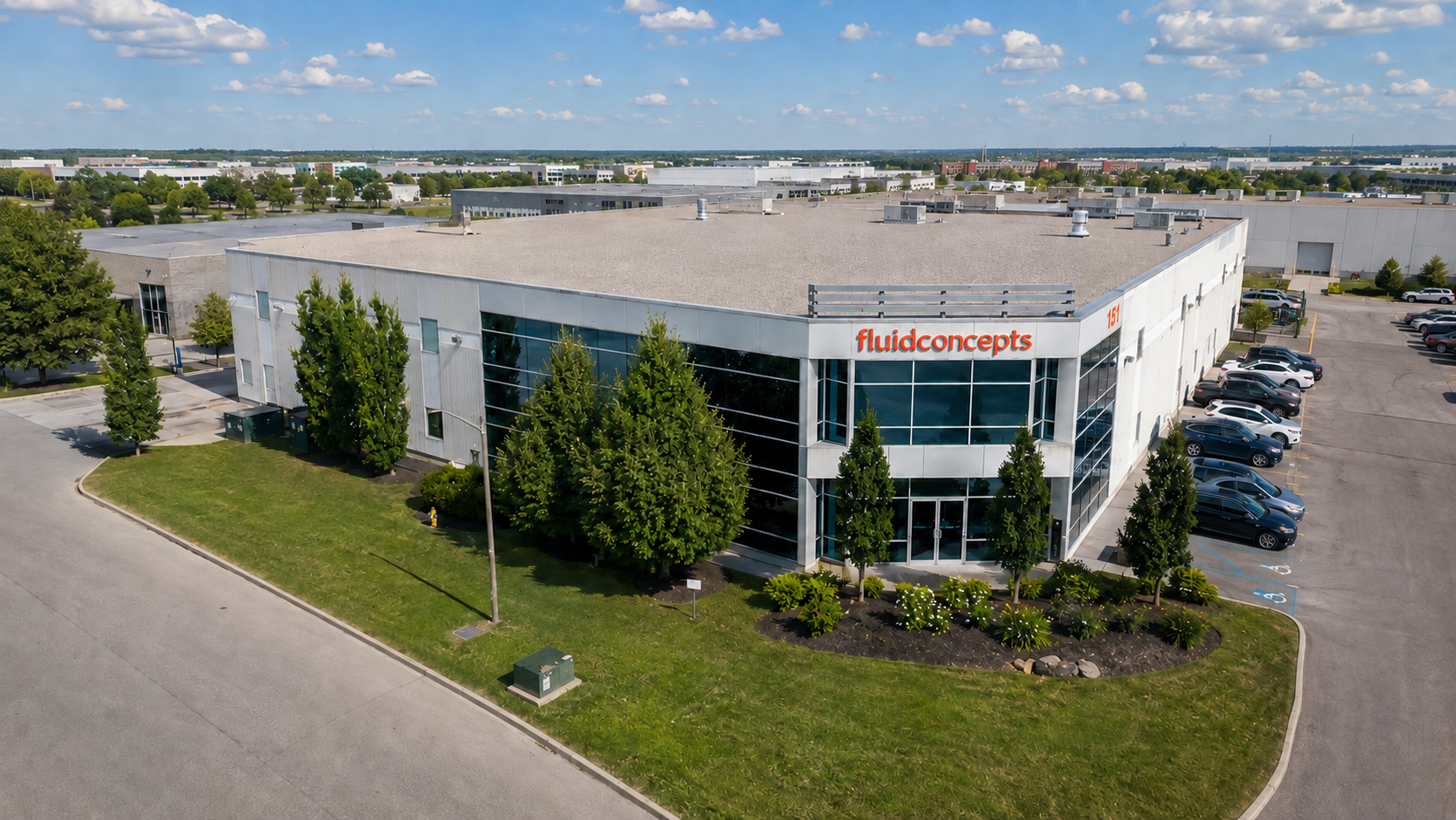 An aerial view of a commercial building with the sign 'fluidconcepts', surrounded by a parking lot with cars and trees, under a partly cloudy sky.