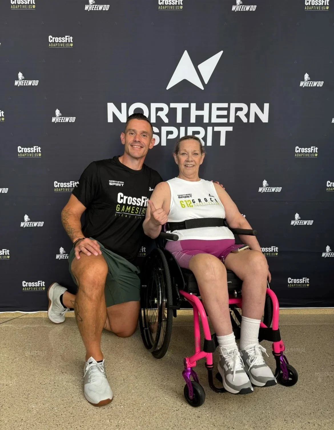 Two people in front of a Northern Spirit backdrop; one is kneeling, and the other is sitting in a wheelchair.