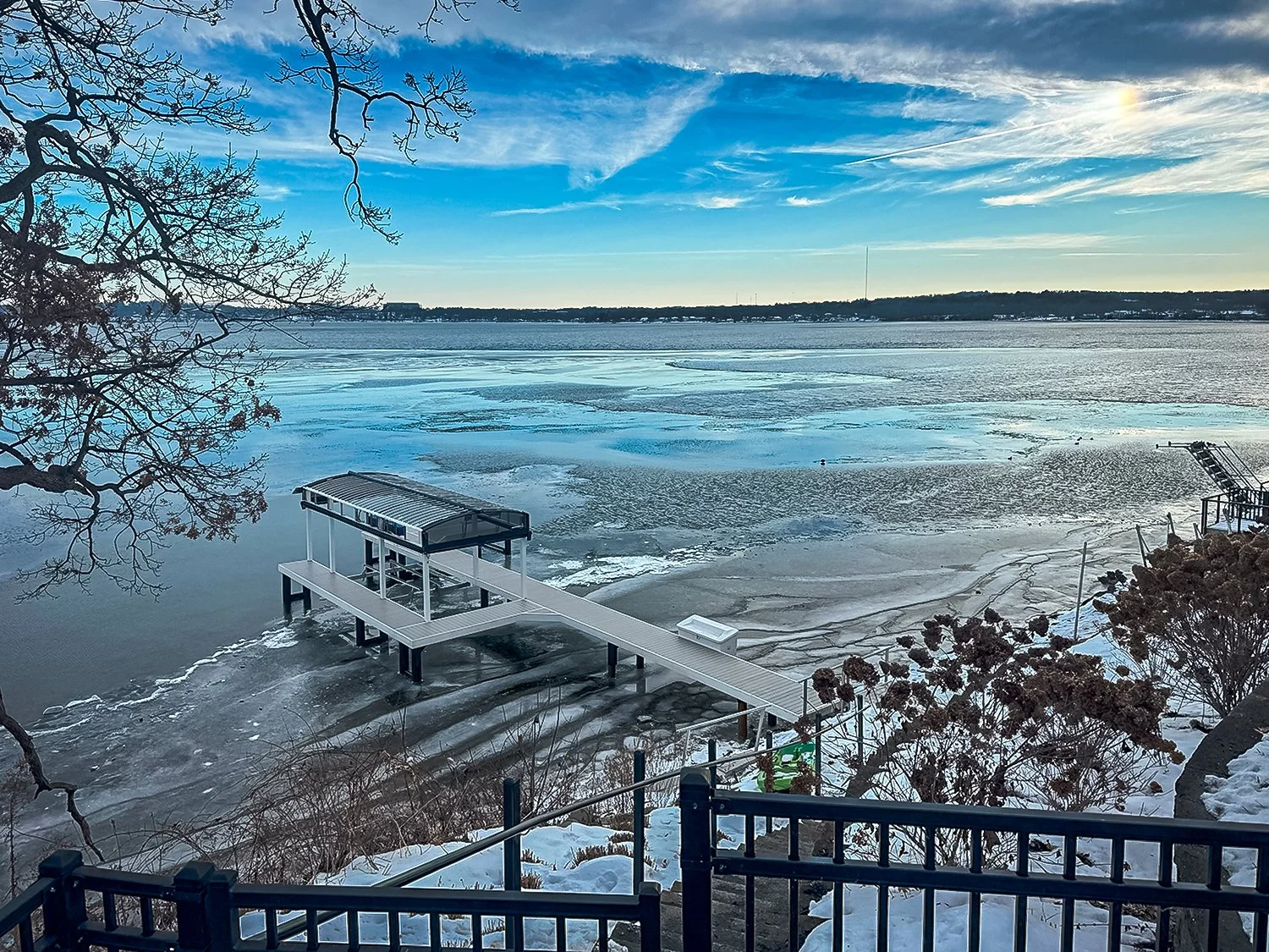 Frozen lake with a dock extending into the ice, surrounded by leafless trees and snow-covered ground, under a blue sky with some clouds.