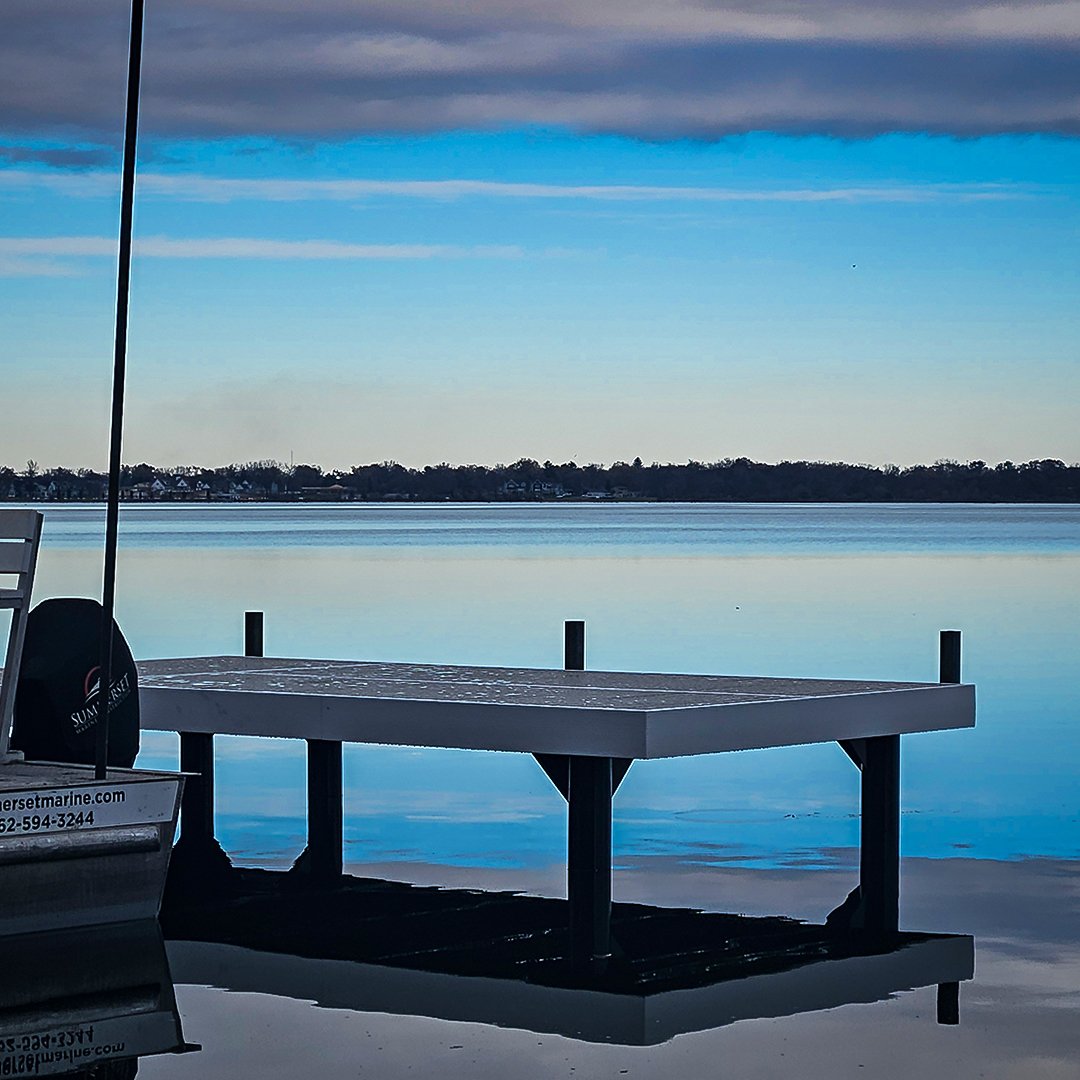 A dock on a calm lake with a partially visible boat on the left and a cloudy sky reflected on the water.