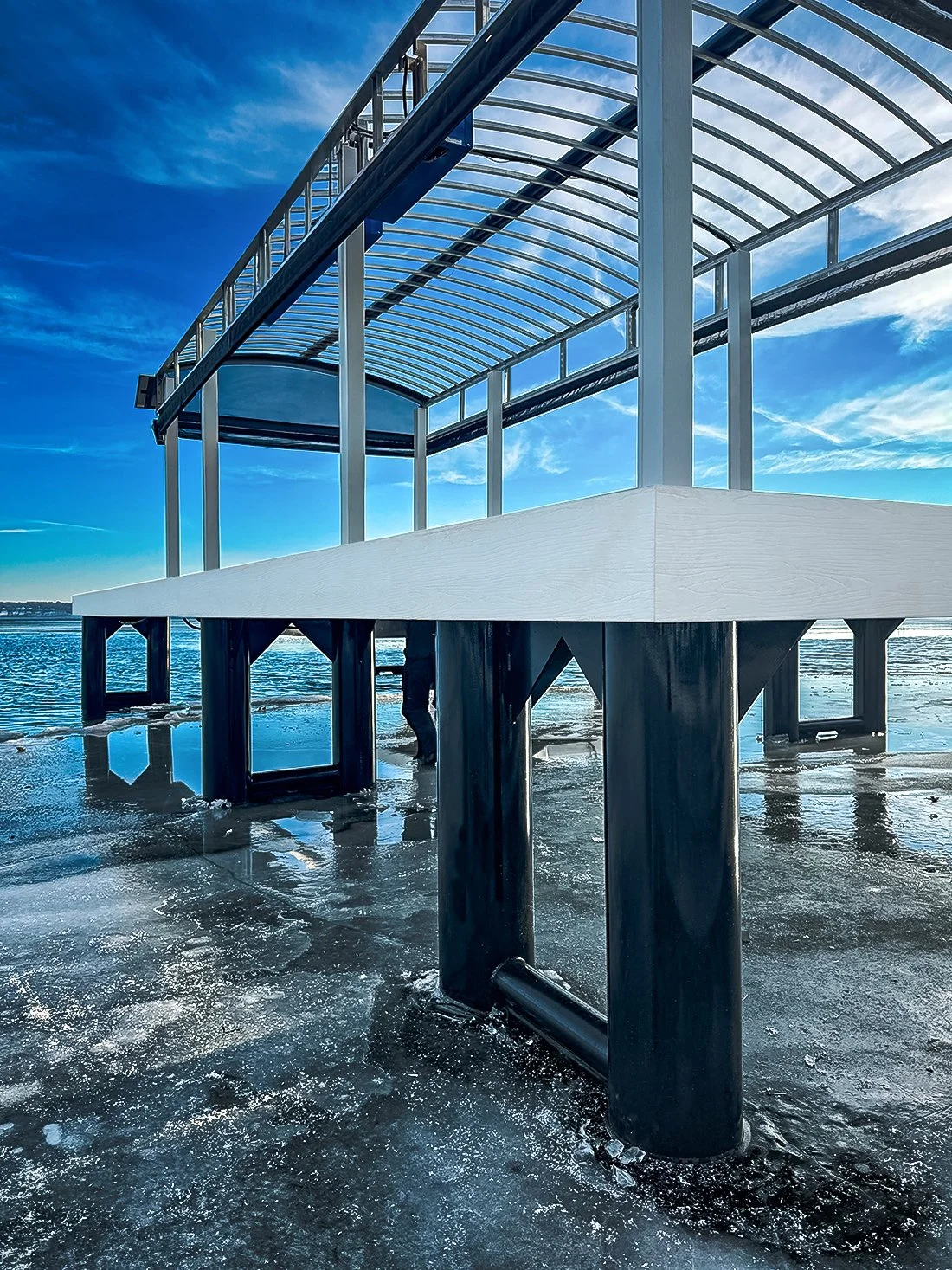 A modern pier or dock extending over frozen water with a clear glass roof structure and metallic supports, beneath a partly cloudy blue sky.