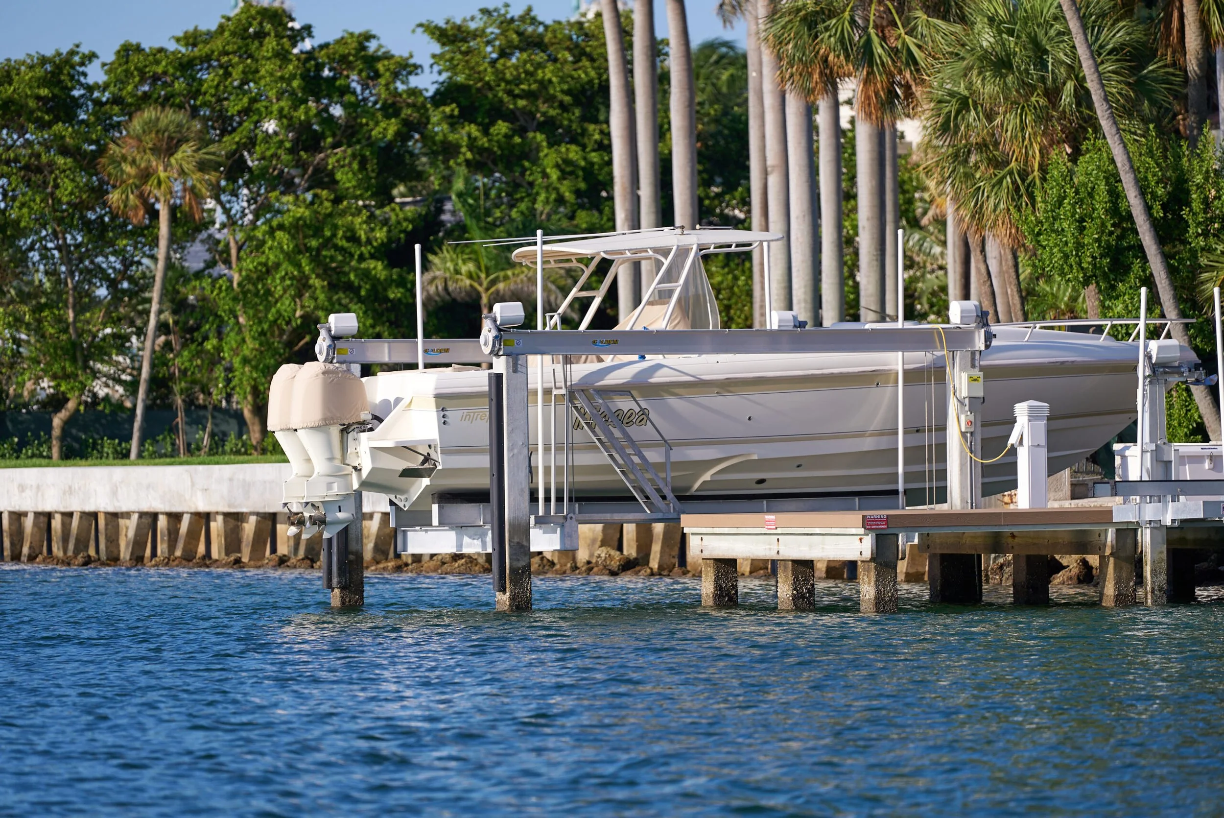 A white Intrepid boat with covered outboard motors is elevated on a boat lift over calm water, with a backdrop of palm trees and a concrete seawall under a clear blue sky..jpg