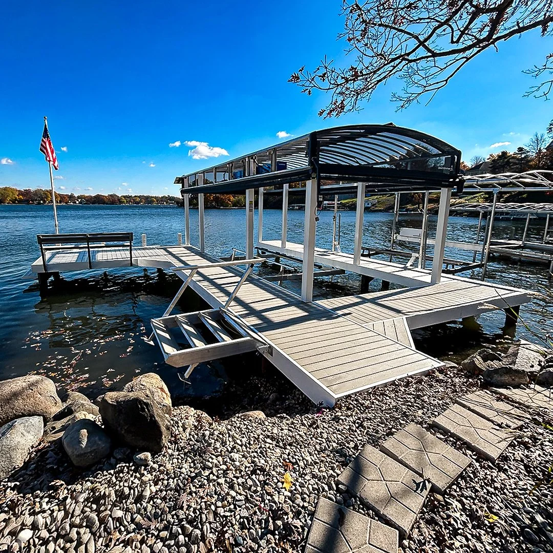 Dock with a covered boat lift on a lake, with rocks and a paved pathway along the shoreline, blue sky, and trees in the background.