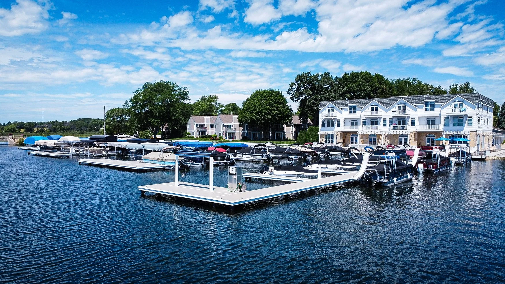 Several boats docked at a marina on a lake with residential buildings and green trees in the background. The sky is partly cloudy, and the water is calm.