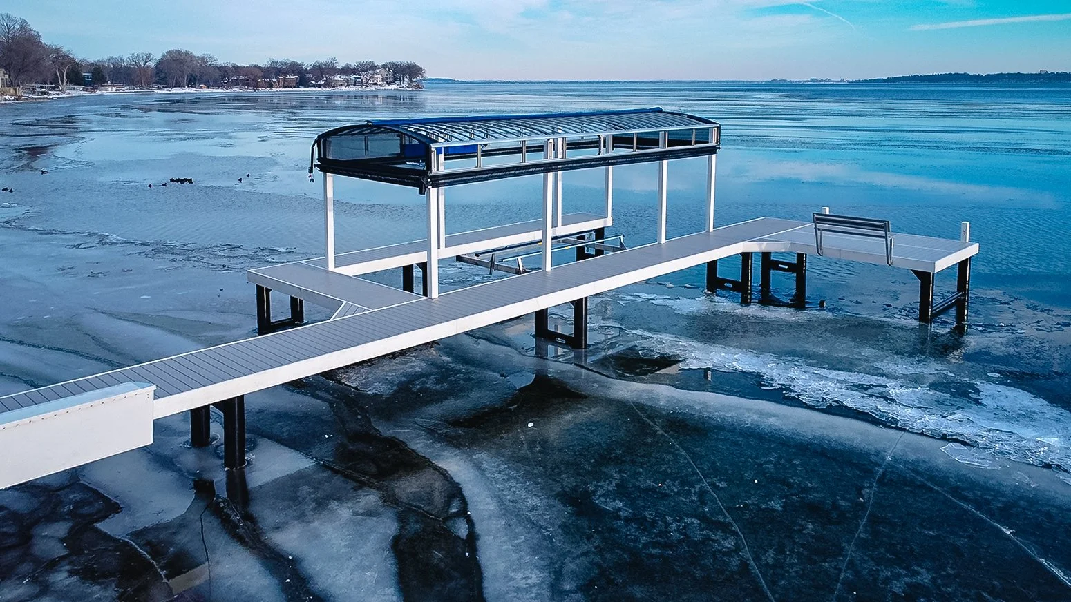 A dock extending over a frozen lake, with a covered platform and benches, surrounded by snow and icy water.