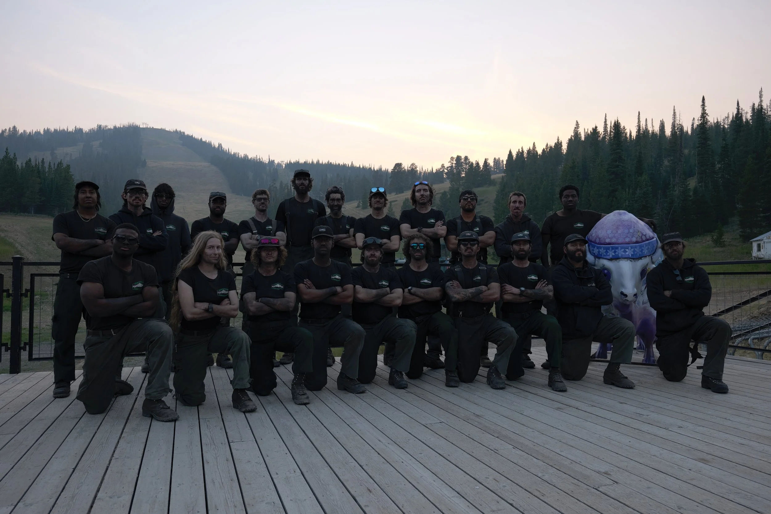 Group of people posing outdoors with mountains and trees in the background, some wearing sunglasses and hats, and a purple inflatable character on the right.
