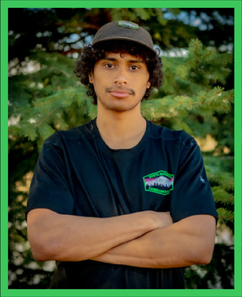 Young man with curly dark hair, wearing a green and gray baseball cap and a black T-shirt with a mountain and forest logo, standing outdoors in front of green trees with arms crossed.