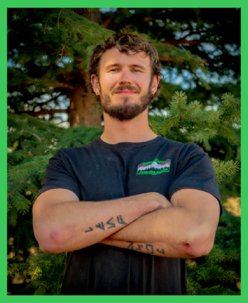 A young man with a beard and curly hair standing with arms crossed in front of a pine tree, wearing a black t-shirt with a patch on the chest.