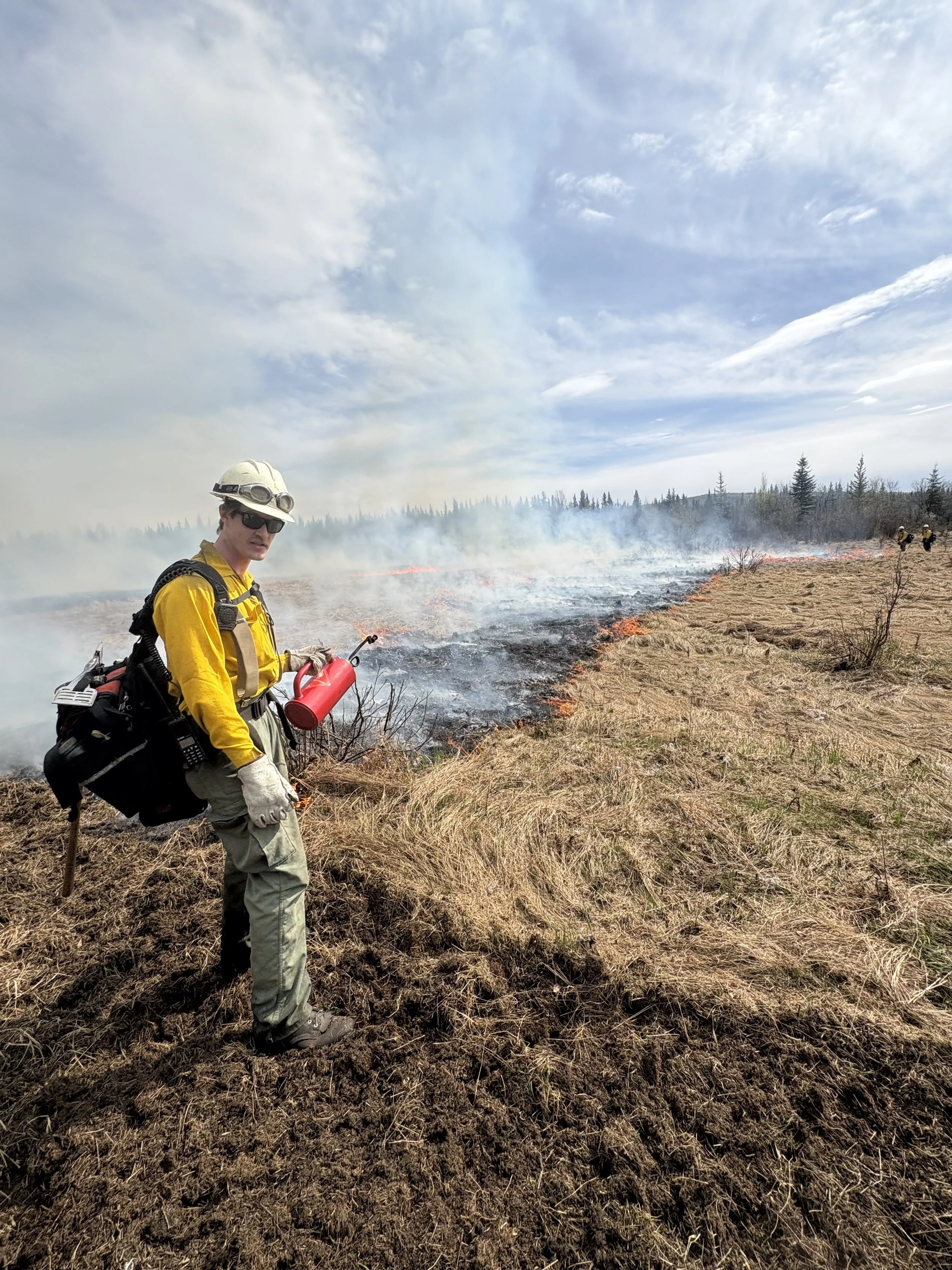 A firefighter stands in a field of dry grass and dirt, holding a "driptorch", with a grass fire burning behind him, and smoke and clouds in the sky.