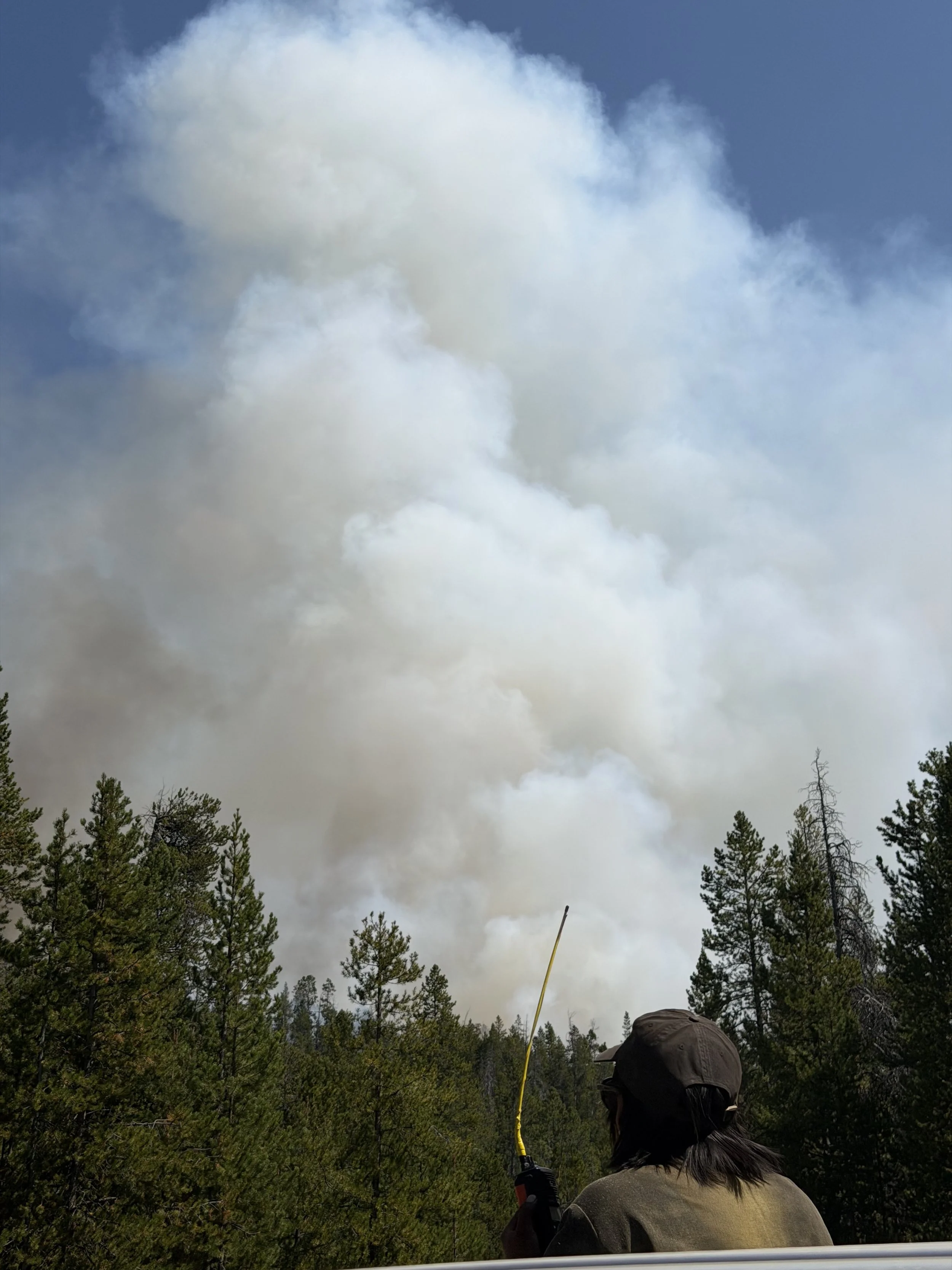 Person with long hair and cap holding a radio looking a big plume of smoke that seems to be close to him.