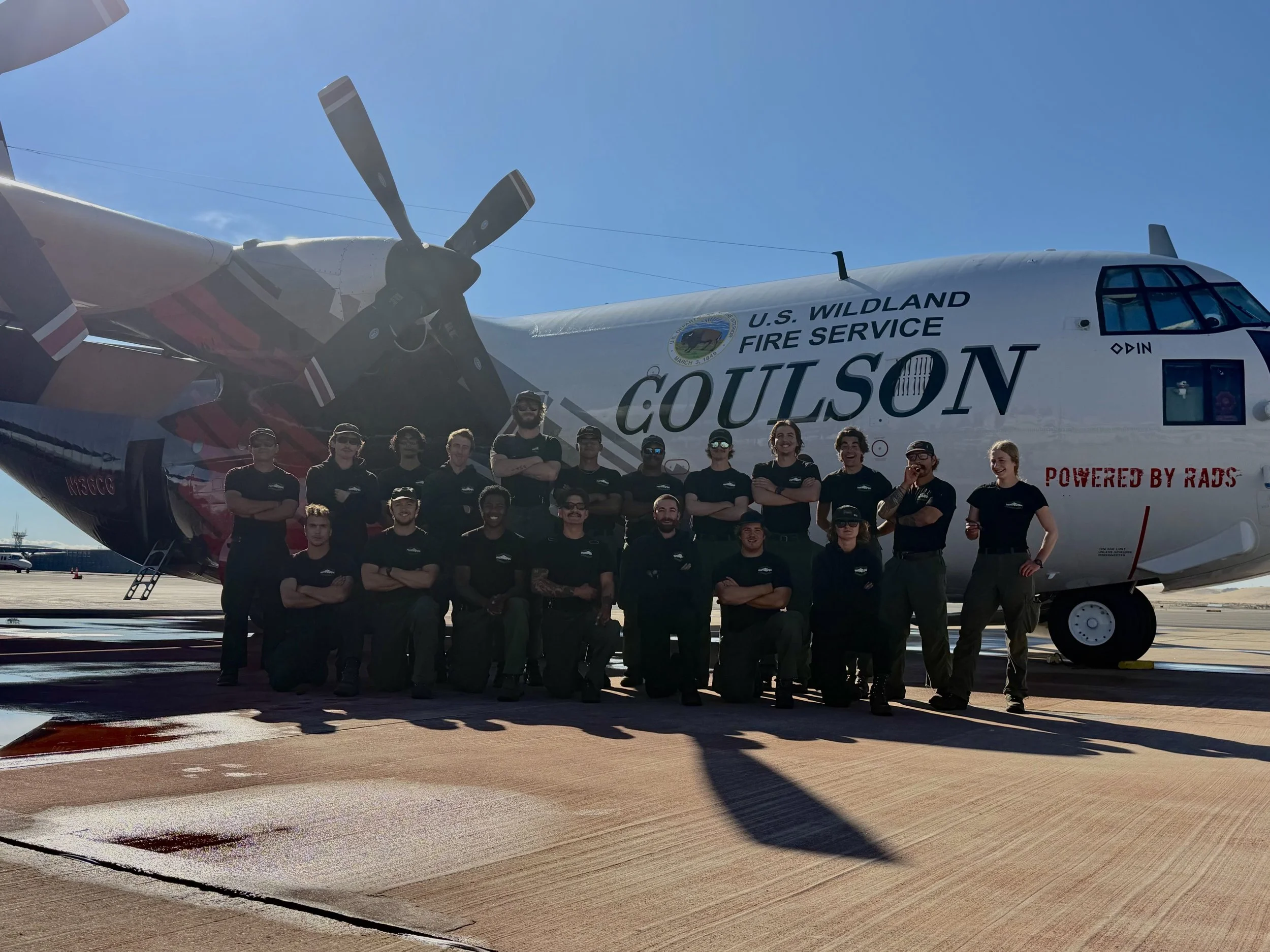 Group of people standing in front of a large airplane with 'Goulson' and 'U.S. Wildland Fire Service' written on it, under a clear blue sky.