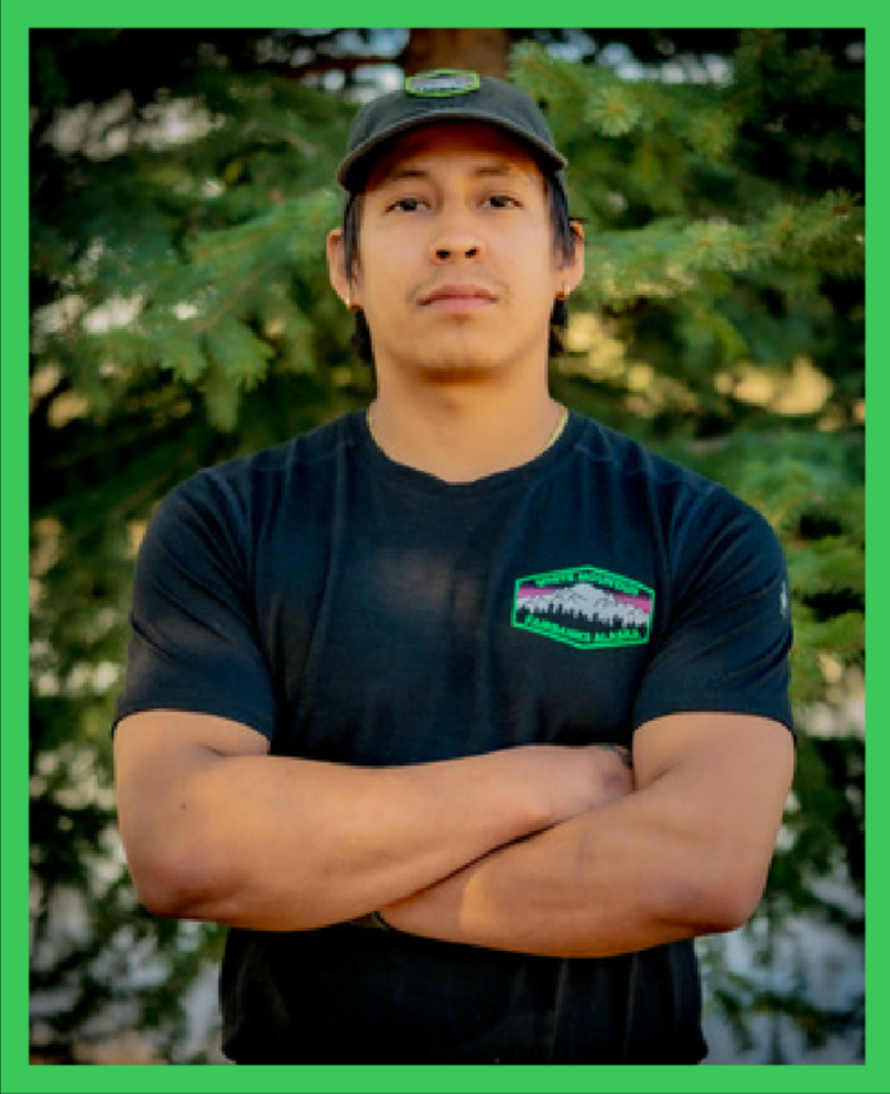 Young man with arms crossed, wearing a black T-shirt with a colorful logo, and a cap, standing outdoors with green trees in the background.