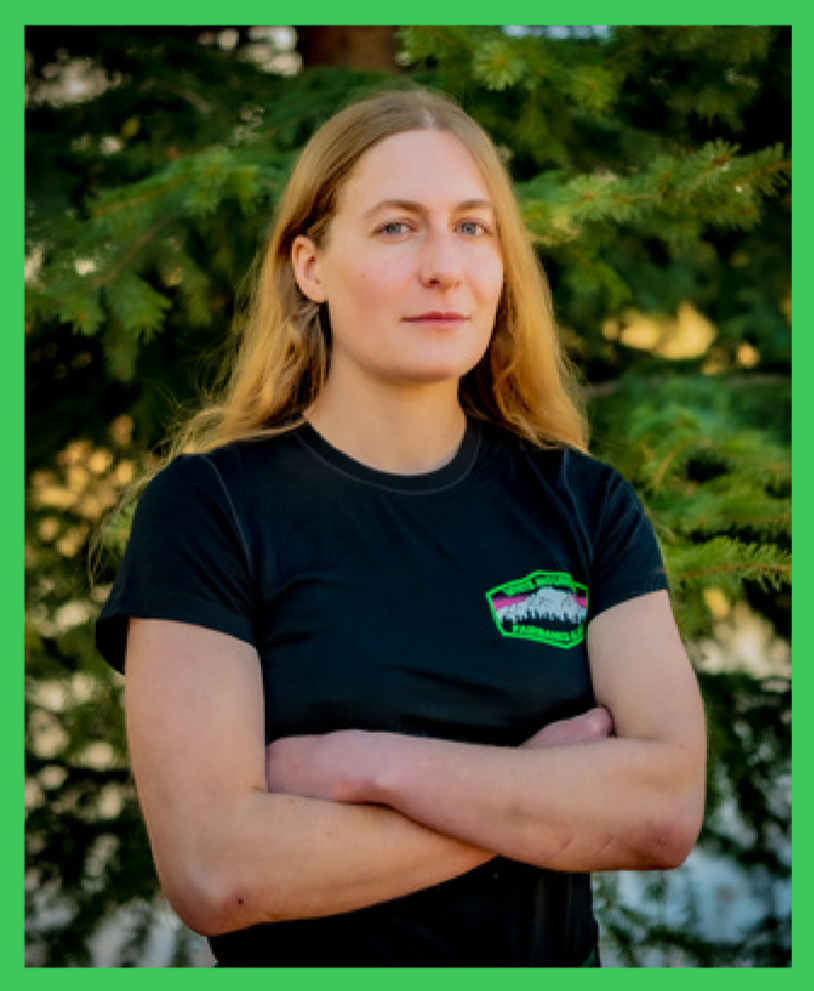 A woman with long blonde hair standing outdoors with crossed arms, wearing a black T-shirt with a White Mountain on the chest, in front of green trees.