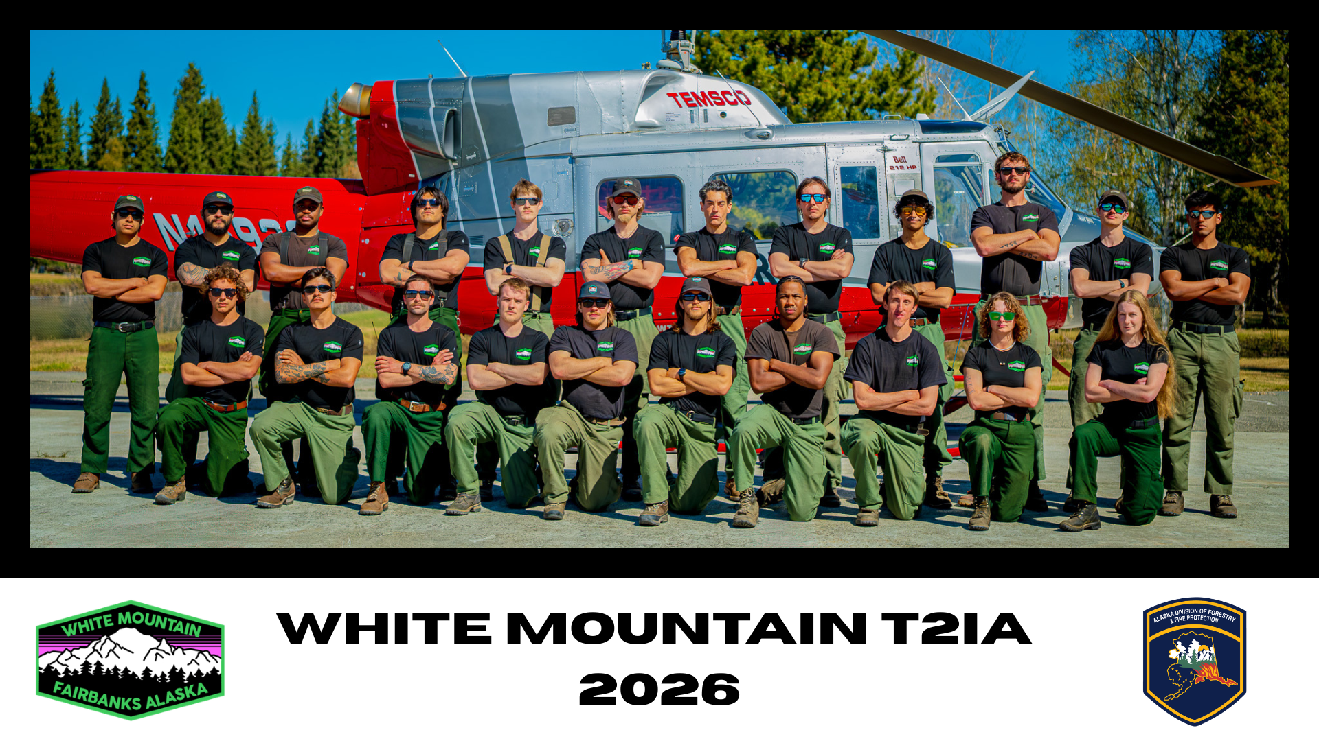 Group of people in uniform standing and kneeling in front of a helicopter with the text 'White Mountain T2IA 2026' and logos for White Mountain Fairbanks Alaska and Alaska Division of Forestry & Fire Protection.