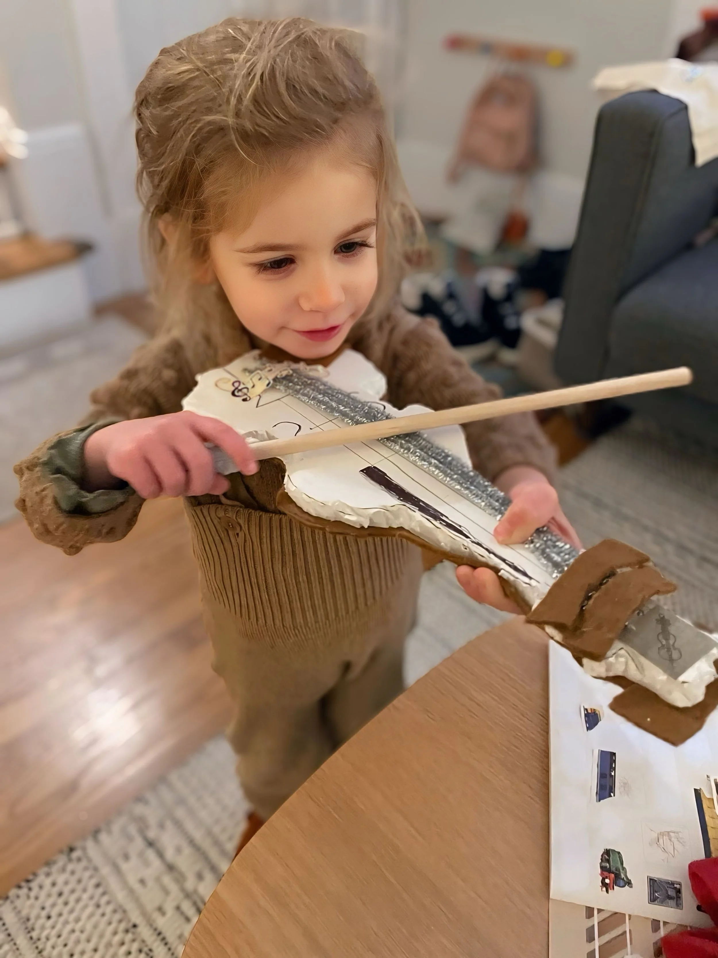 Exploring music through play — Kleo practices with her cardboard violin and piano at home, bundled up and smiling. 