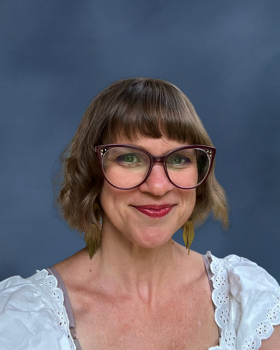 Portrait of a woman with short brown hair, glasses, a pearl necklace, and a floral jacket, smiling in front of a blue background.