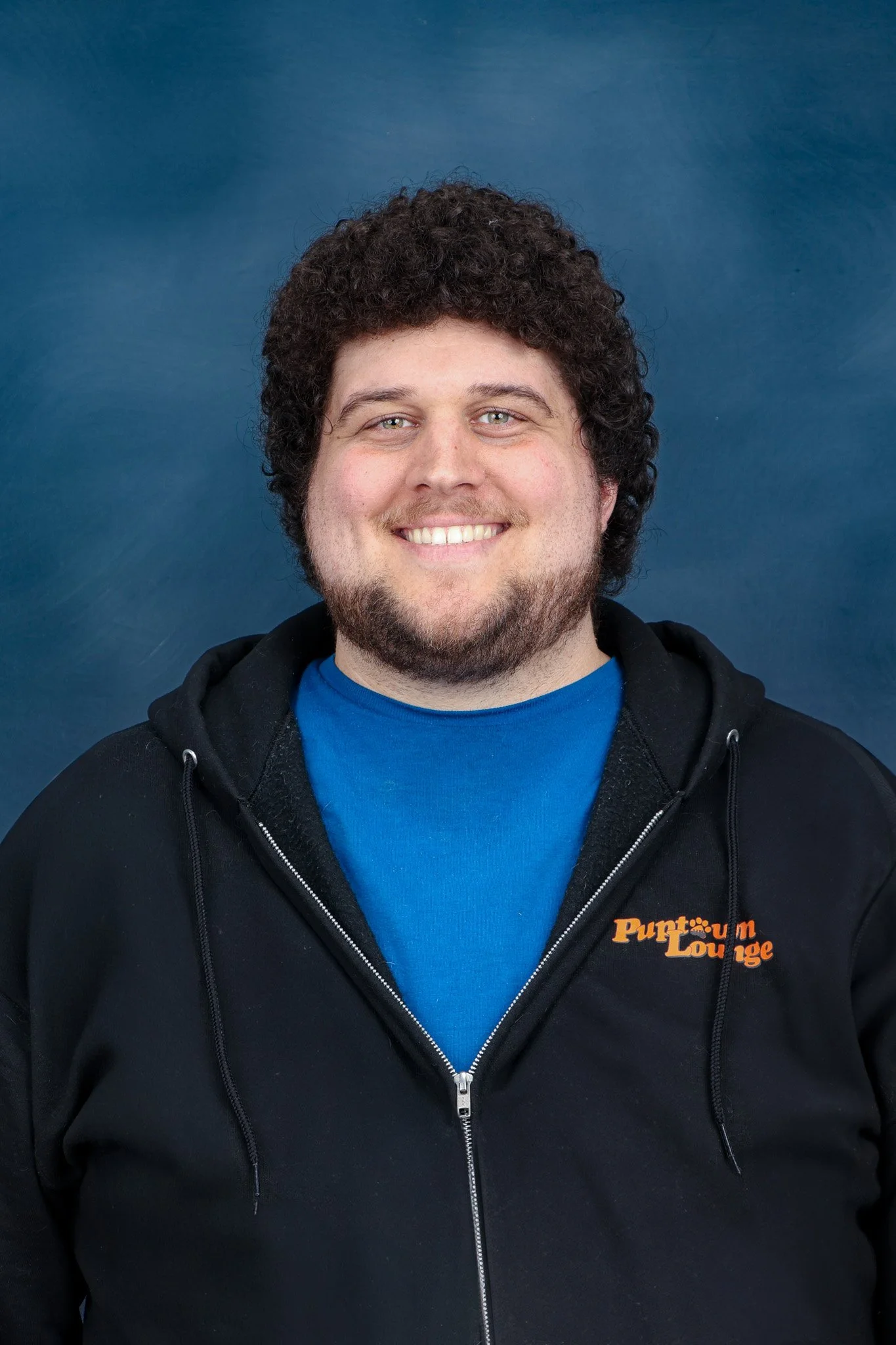 Portrait of a young man with curly black hair wearing a Puptown Lounge Black Hoodie.