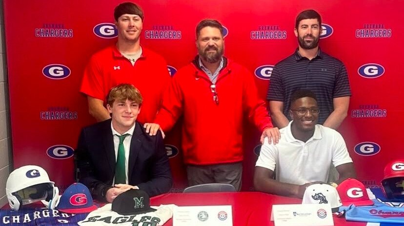 Two students sitting at a table with helmets and sports caps, wearing formal attire, with three adults standing behind them, all in front of a backdrop featuring the logo of a sports team, "Chargers."