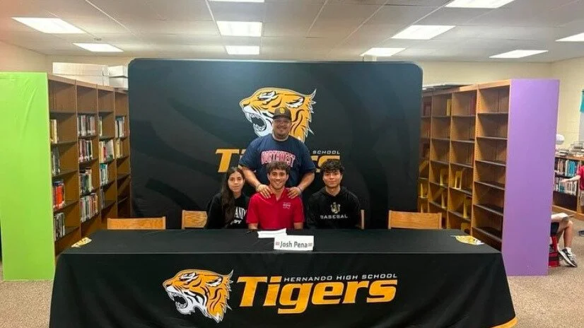 Group of four people posing in a library with a Hernando High School "Tigers" backdrop. One person is standing behind three people sitting at a table with a name card that says "Josh Pena." Shelves with books are visible in the background.