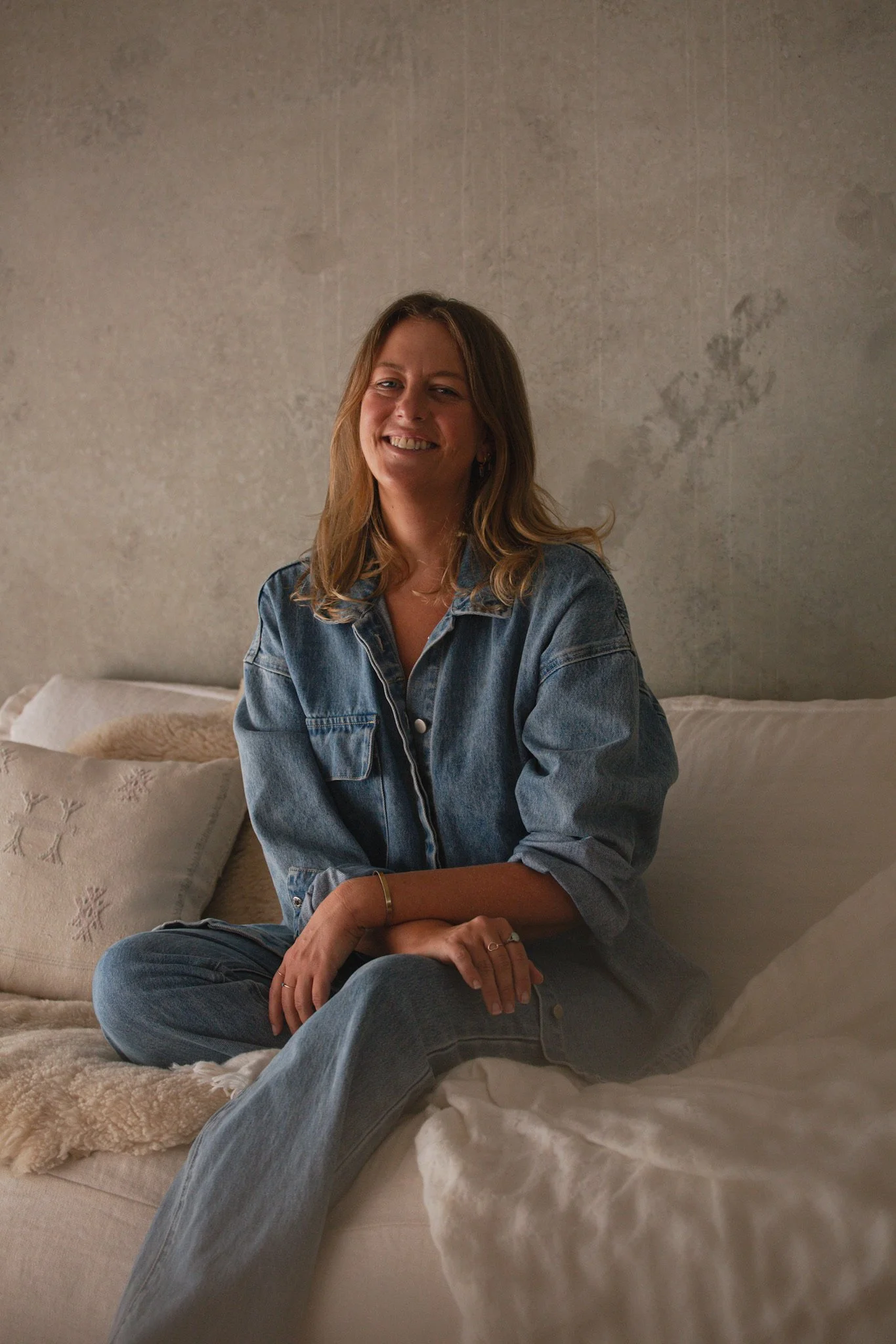 A woman sitting cross-legged on a bed, smiling, wearing a denim jacket and jeans, with pillows and blankets in the background.