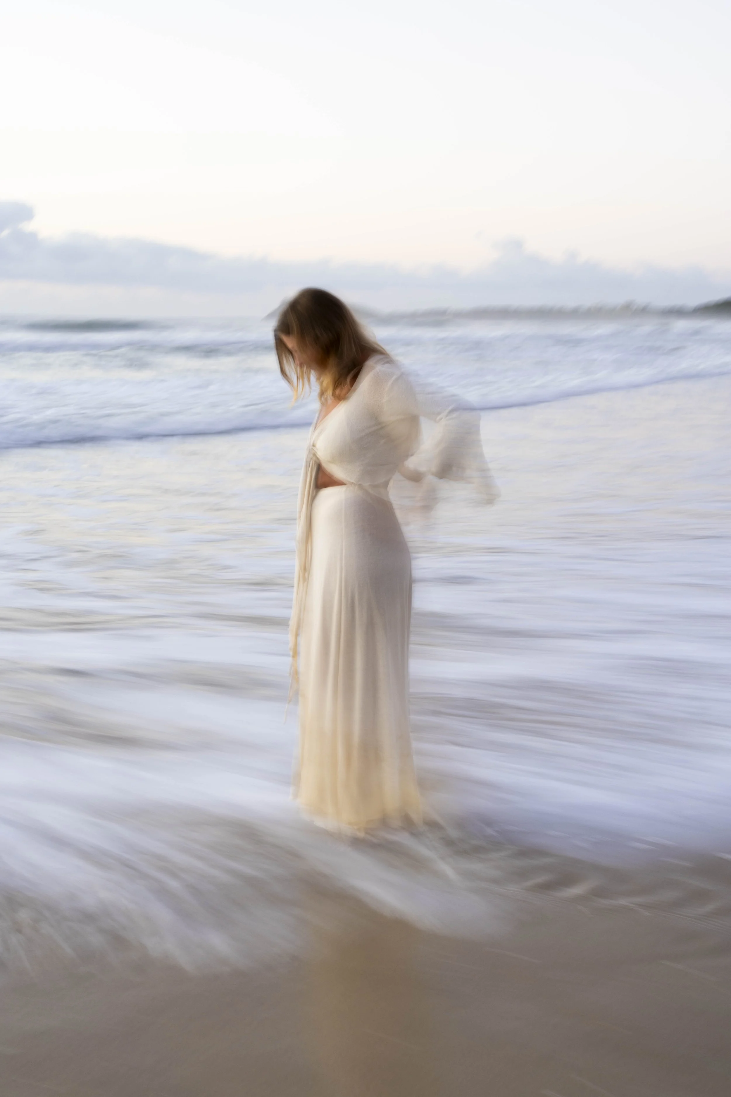 A woman in a white dress standing in the ocean waves during sunset or sunrise.