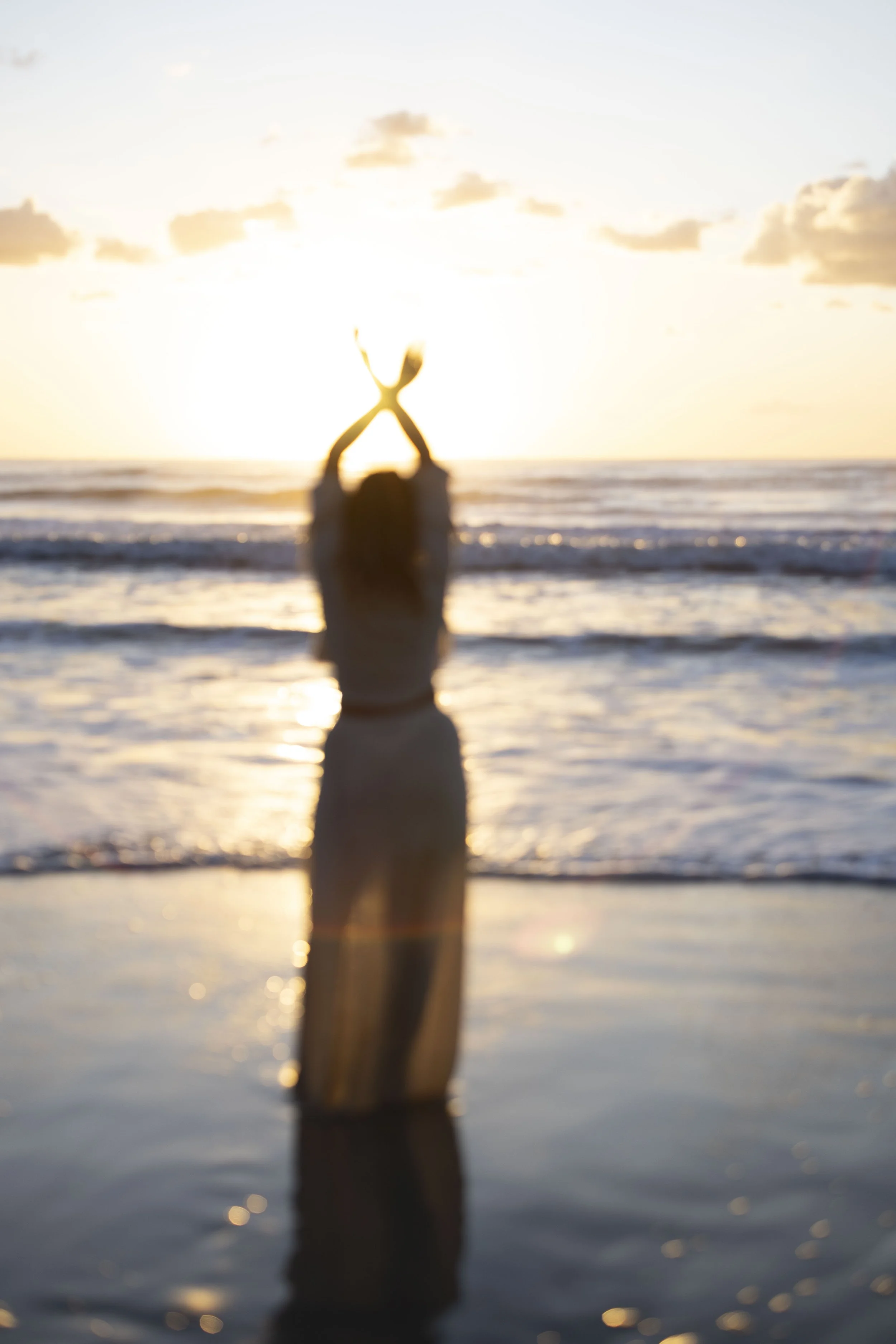 Silhouette of a woman standing in the water at the beach during sunset, with her arms crossed above her head, and the sun setting behind her.