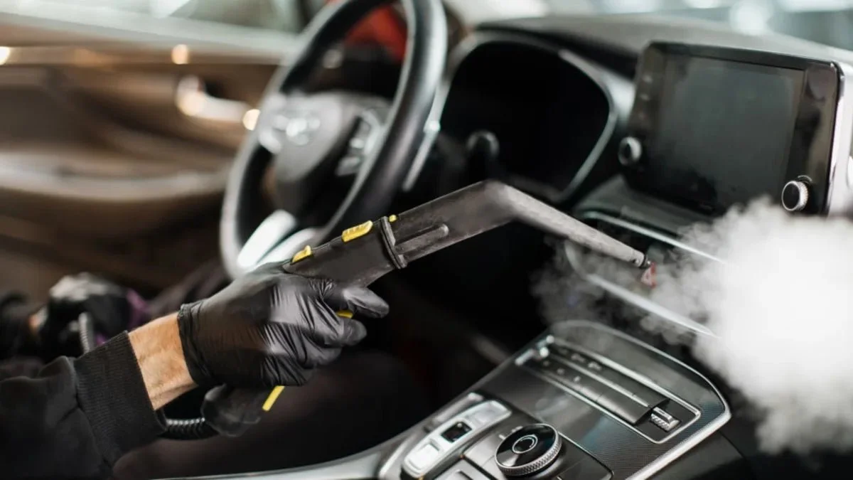 Cleaning car interior with a steam cleaner; steering wheel and dashboard visible.