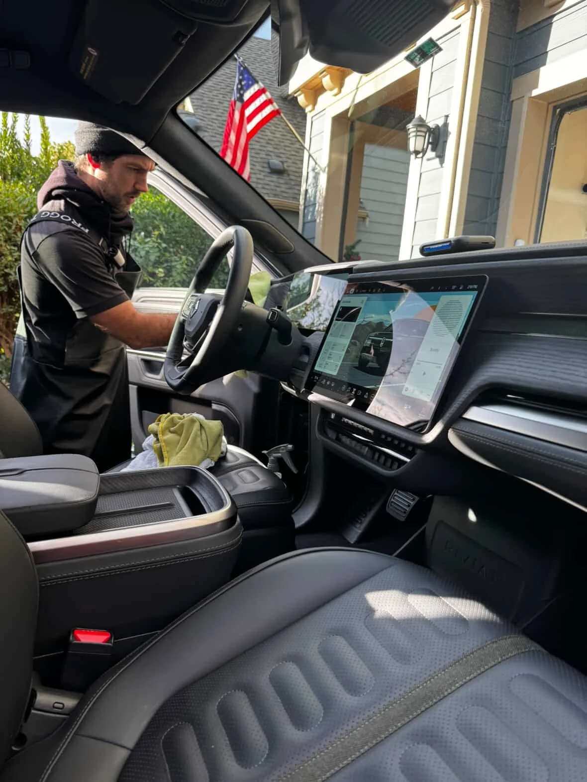 Man in black shirt with a beanie hat standing inside a vehicle, working on something near the dashboard in front of an American flag outside a house with light-colored siding.