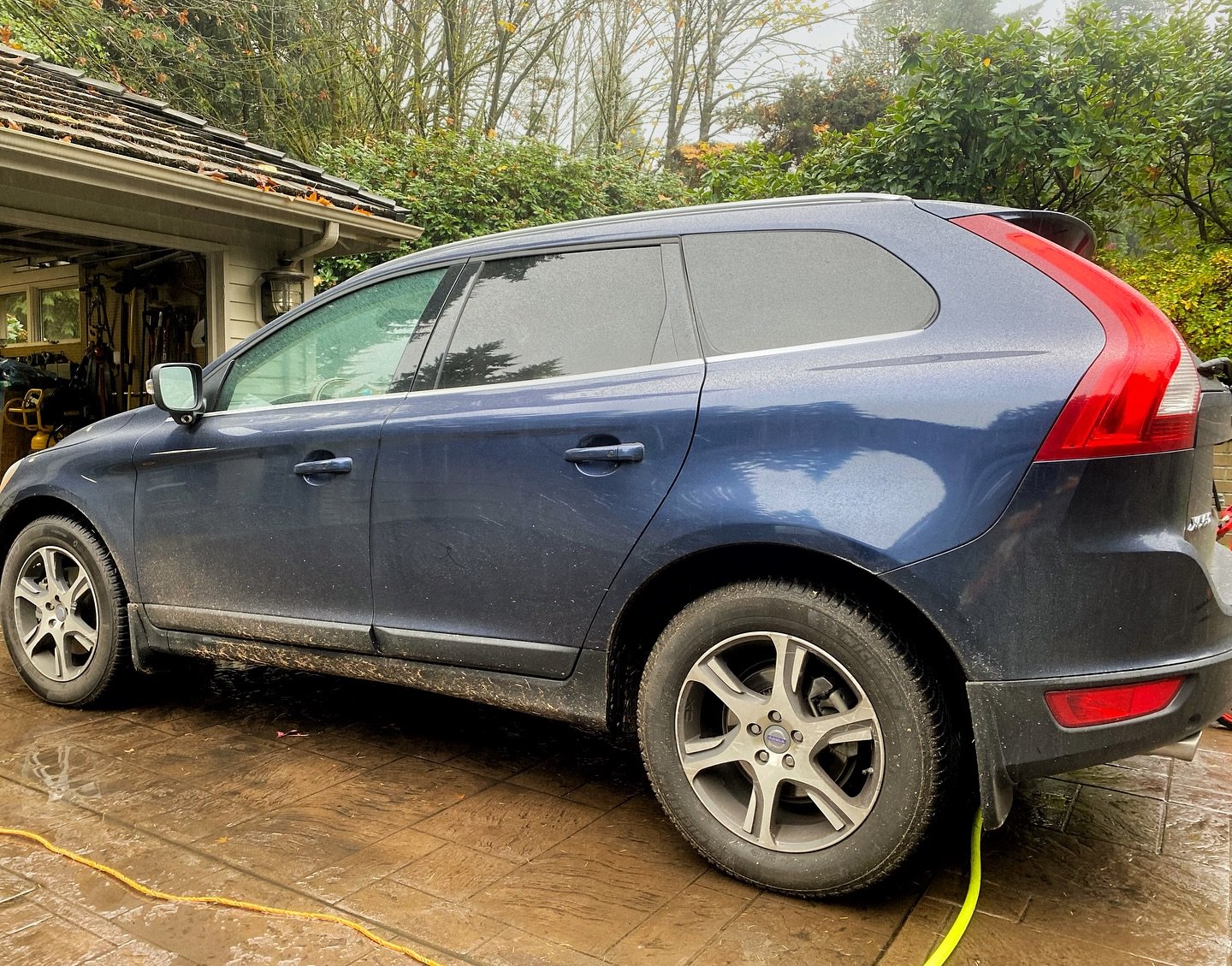 A blue Volvo hatchback parked on a wet driveway in front of a garage, with muddy tires and a yellow gardening hose on the ground.