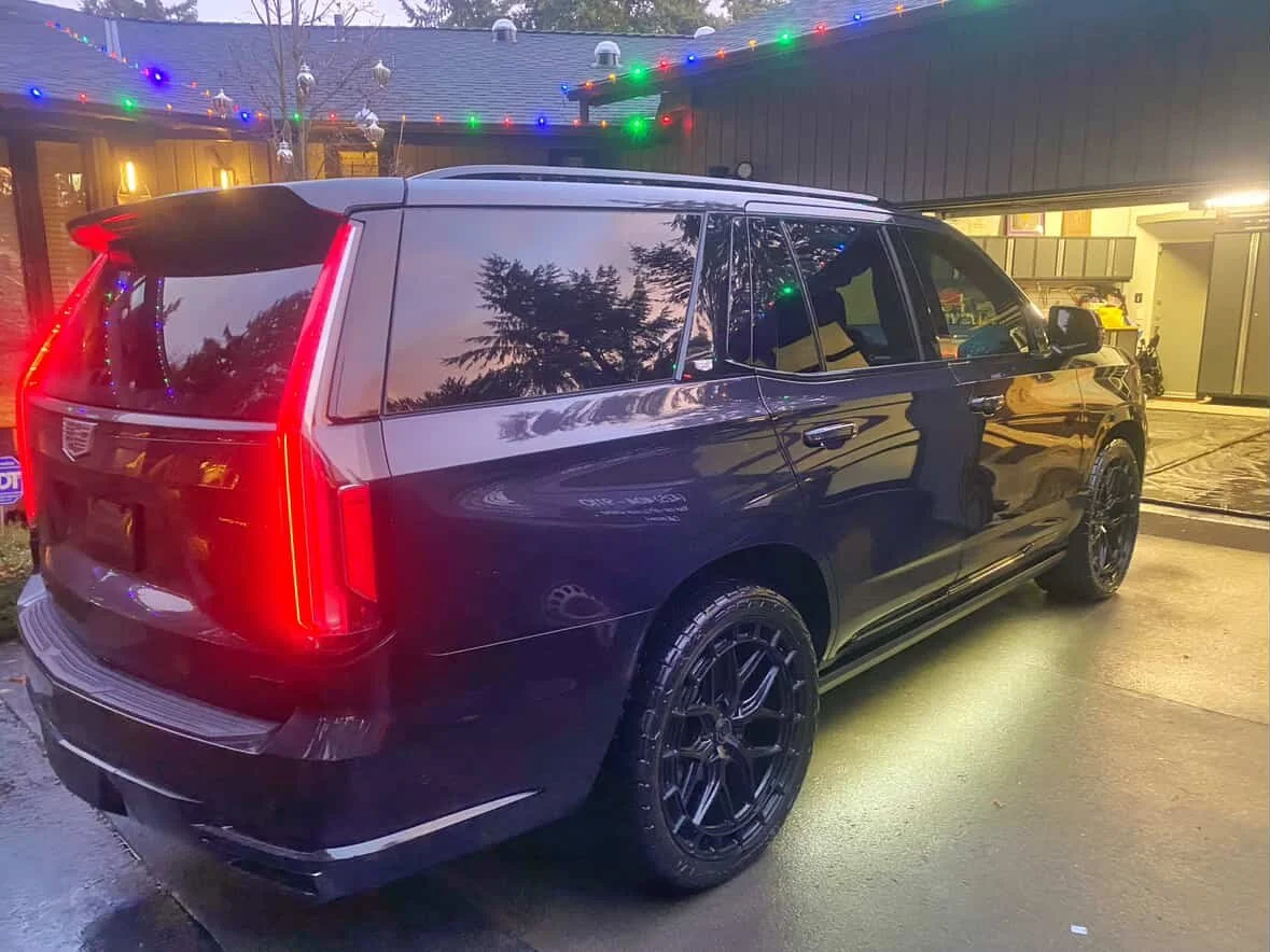 Black Cadillac SUV parked outside house decorated with multicolored string lights during evening or dusk.