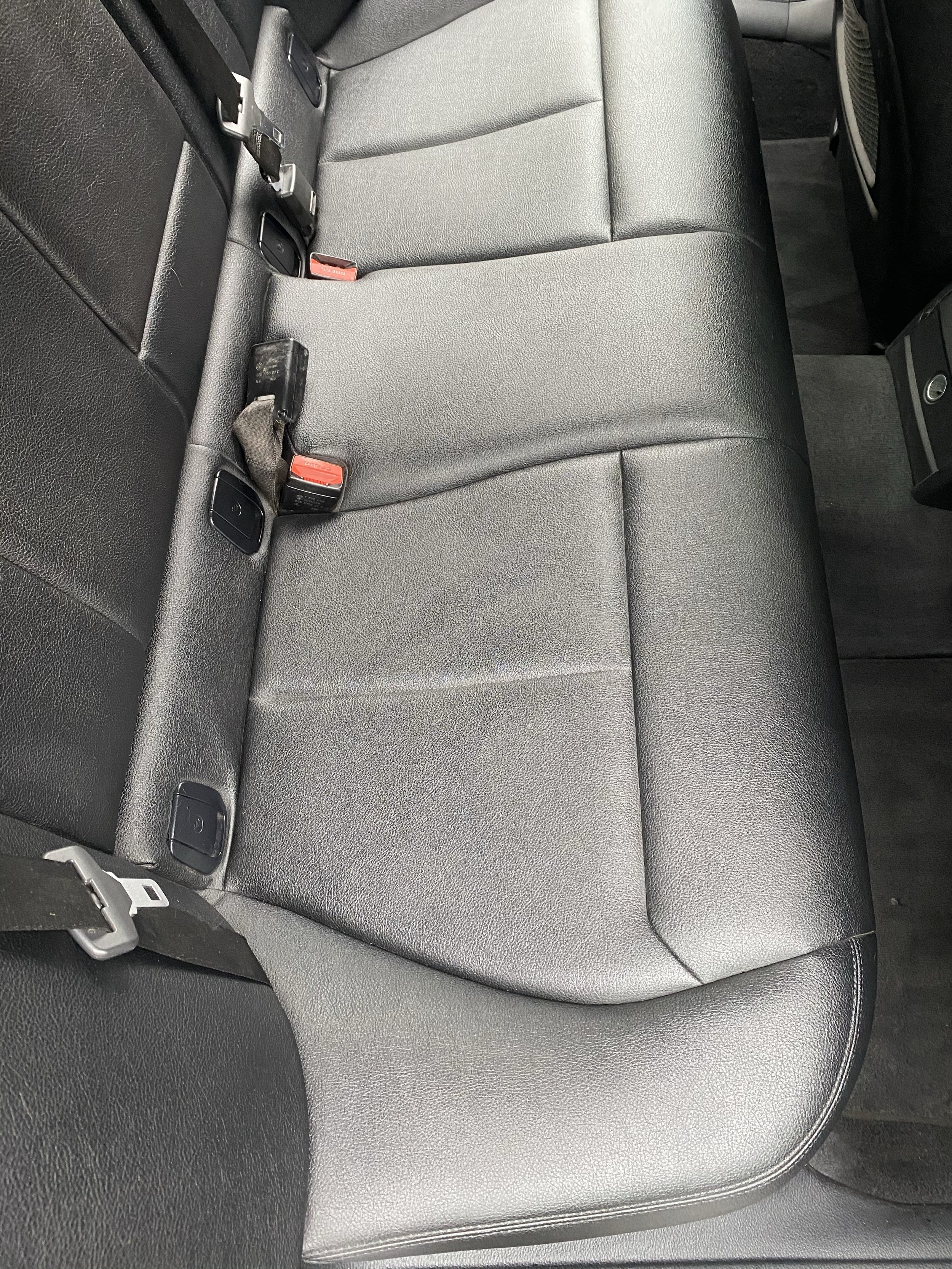 The back seat of a car with black leather upholstery, seat belt buckles, and seat belt straps.